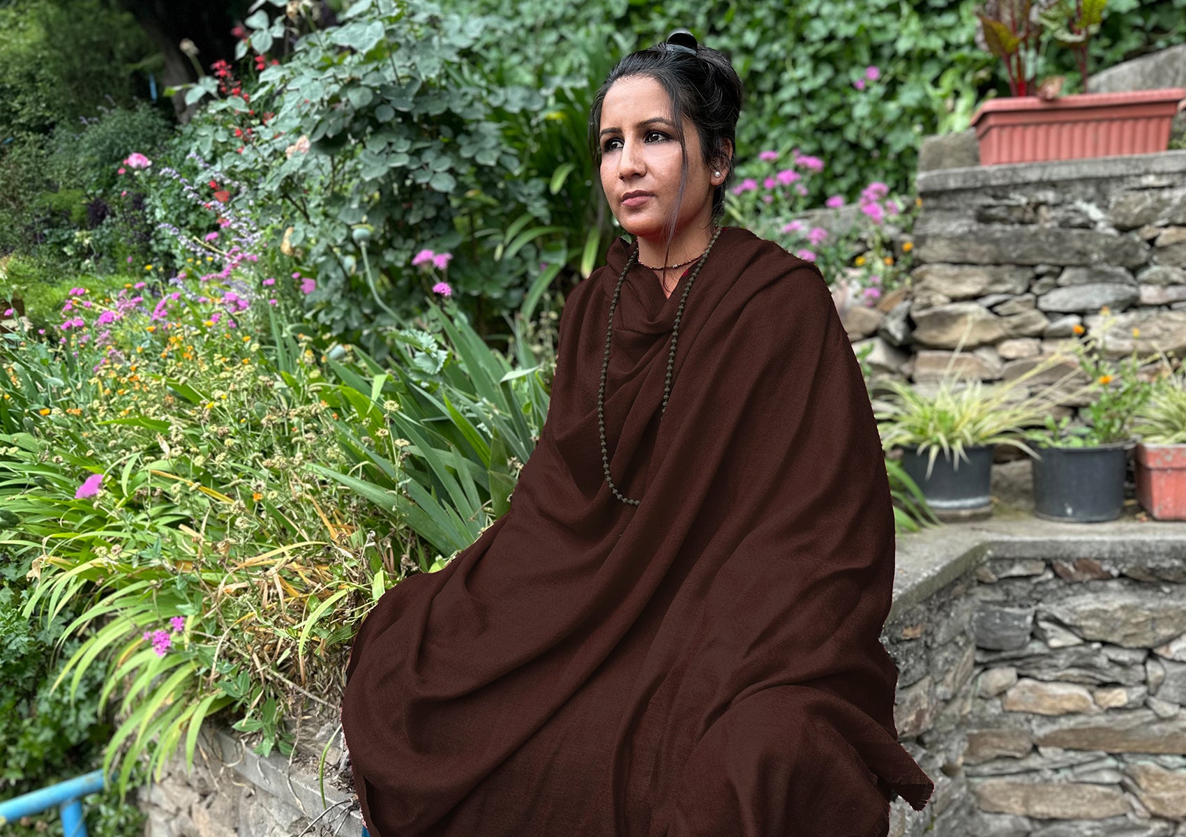 A woman in a brown shawl standing outdoors, with greenery and colorful flowers in the background.