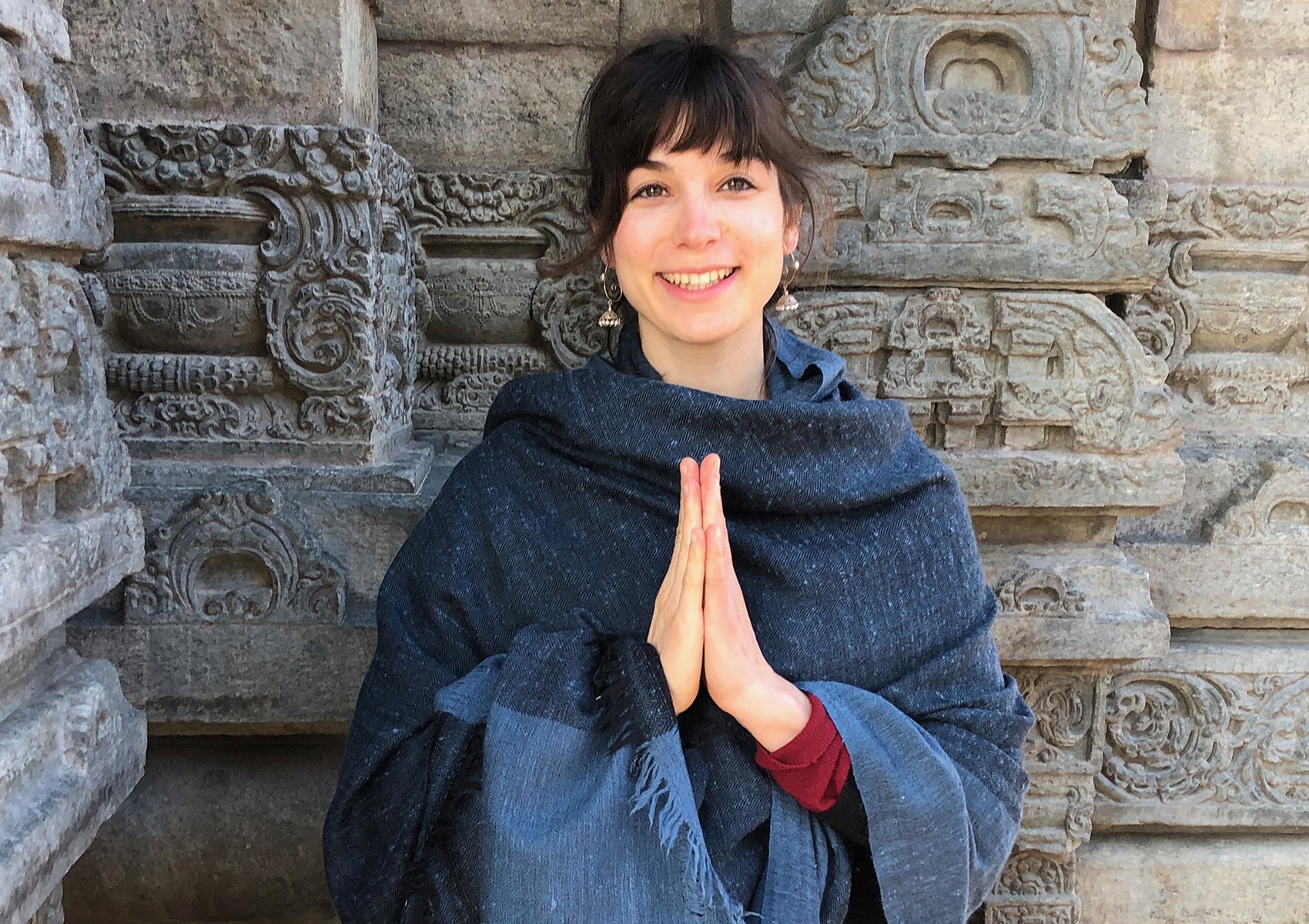 Woman wearing a blue shawl, standing near an intricately carved stone wall.