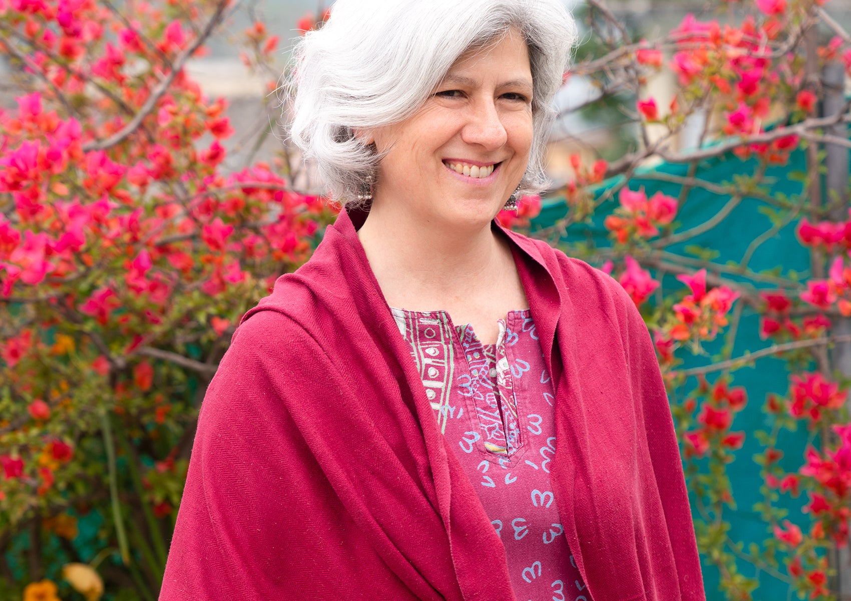 Mature woman in a burgundy shawl against a backdrop of pink flowers, smiling warmly.