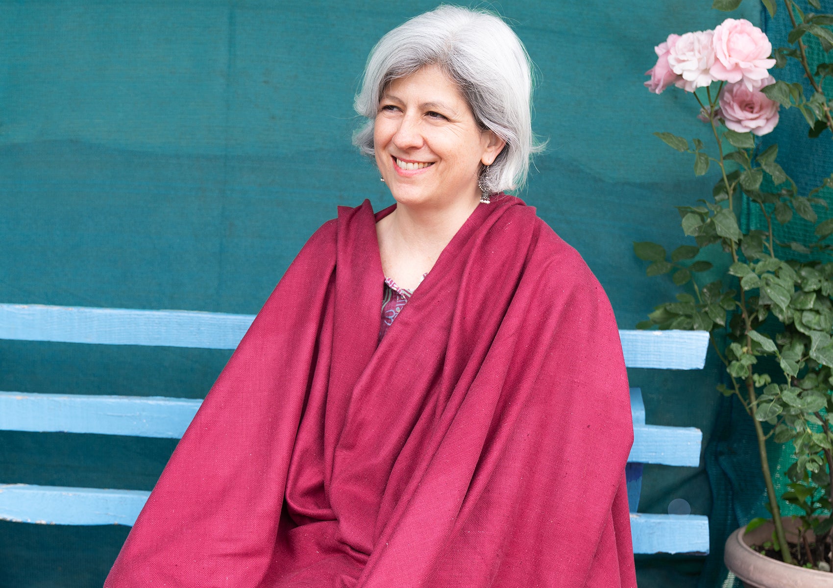 Older woman sitting contently in a burgundy shawl, with a blue wooden bench and potted plants in the background.