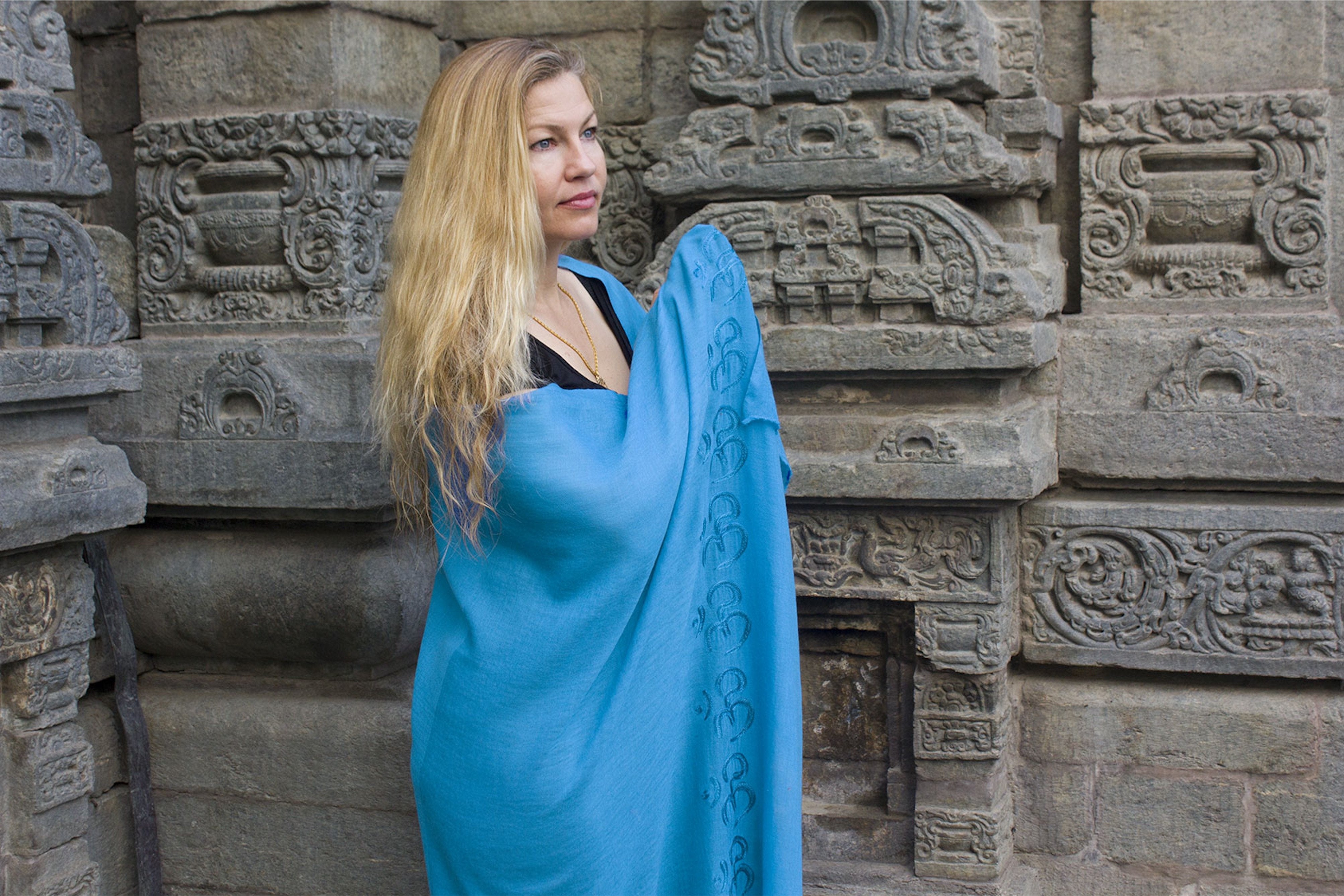 A blue meditation shawl featuring 'Om' symbols, draped over the shoulders of a person standing by ancient stone carvings.