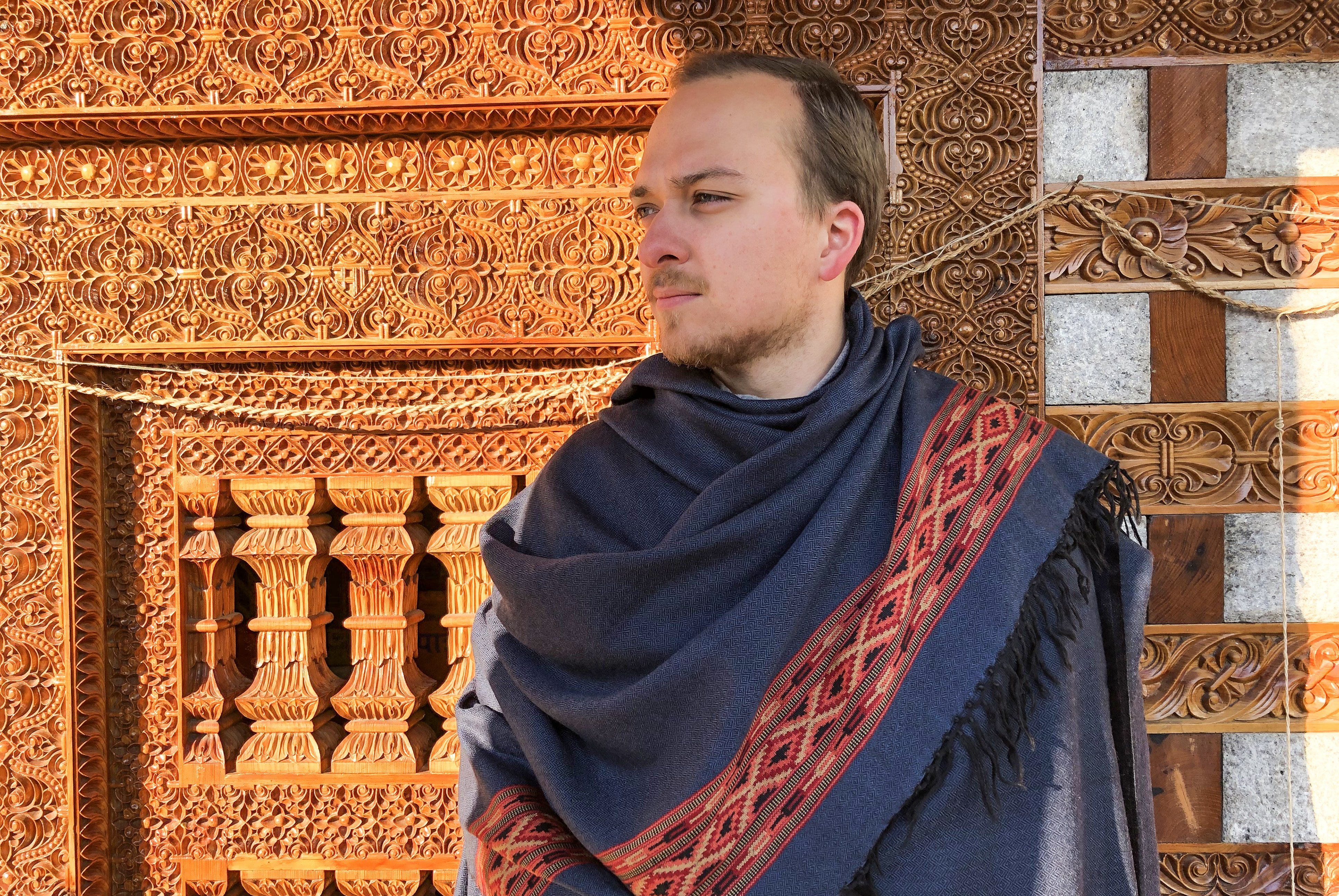 Male wearing a blue meditation shawl in an ancient garden setting with carved stone architecture.