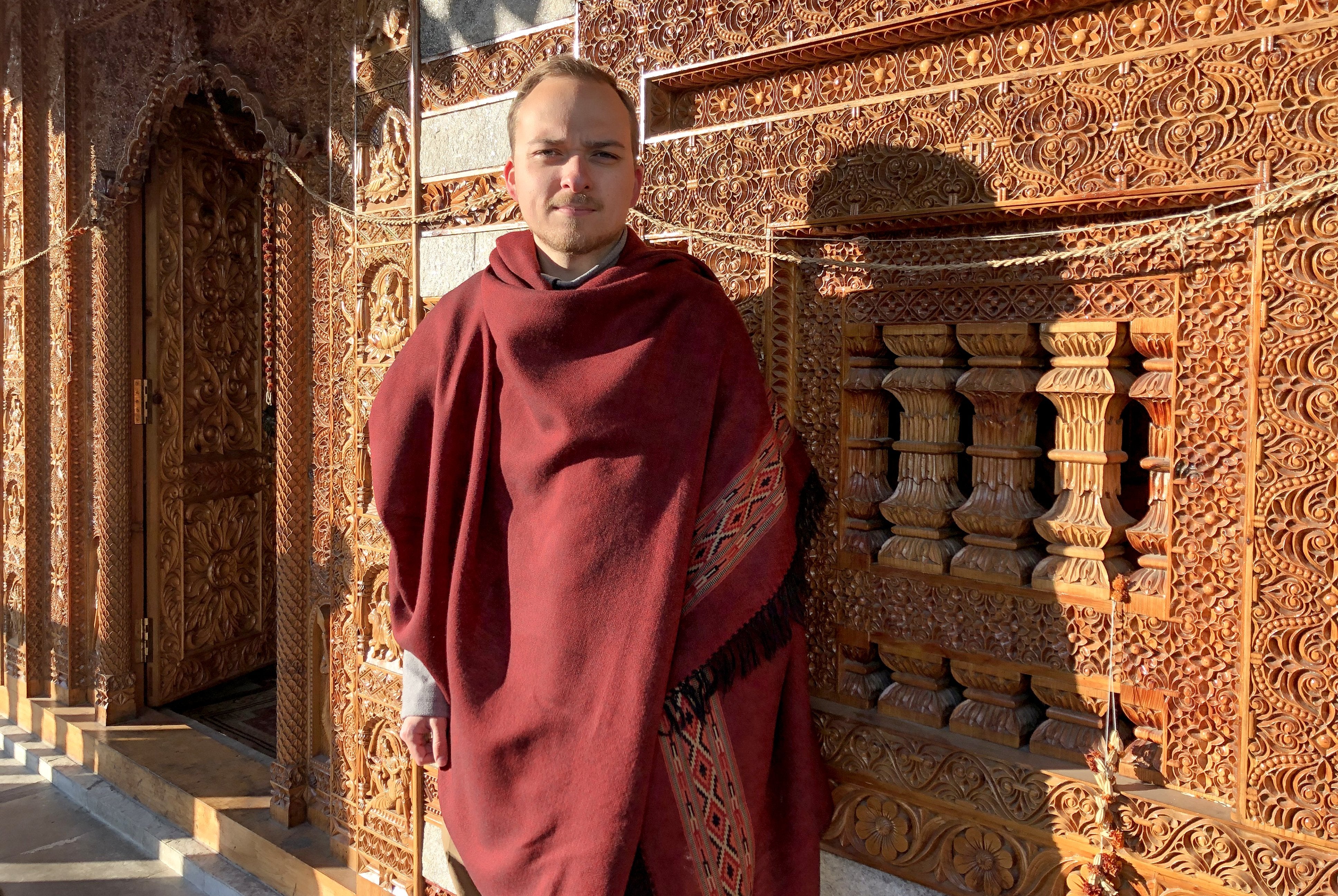 Person wearing a burgundy red meditation shawl with intricate patterns, standing against detailed wooden architecture.