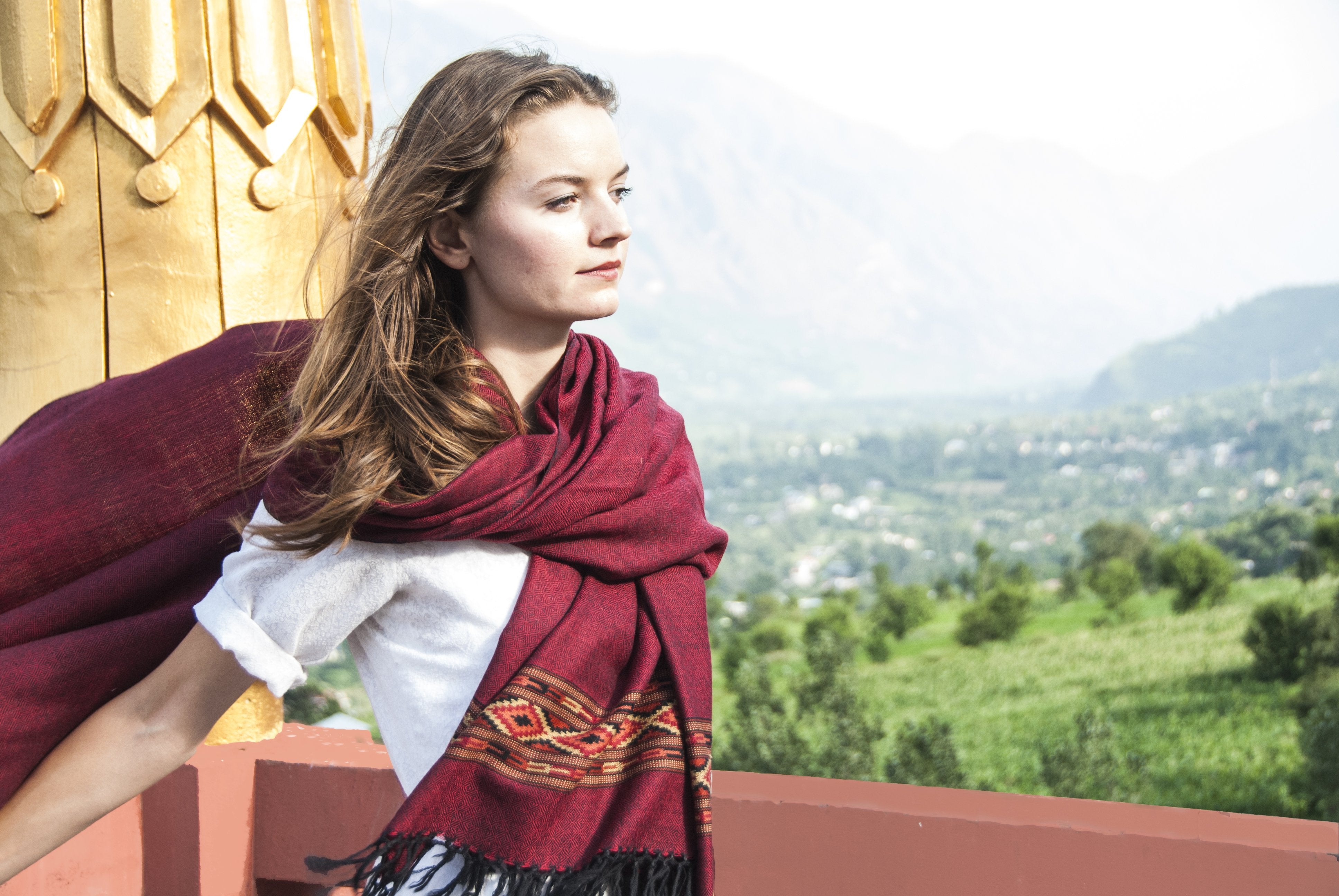 Young woman with a burgundy red meditation shawl wrapped around, looking into the distance with a mountainous view behind.