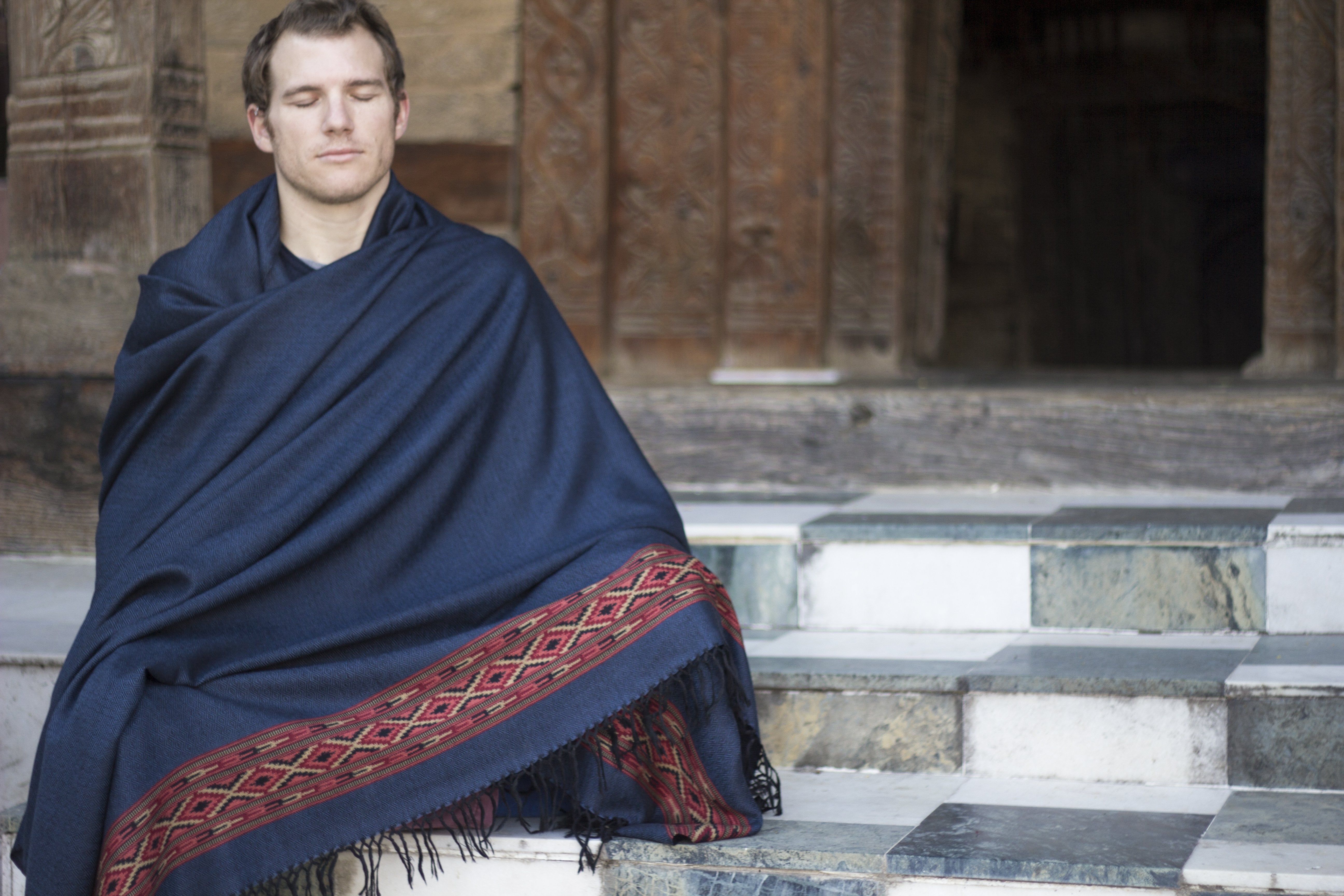 Man sitting in meditation with a dark blue meditation shawl featuring ethnic patterns, on marble temple steps.