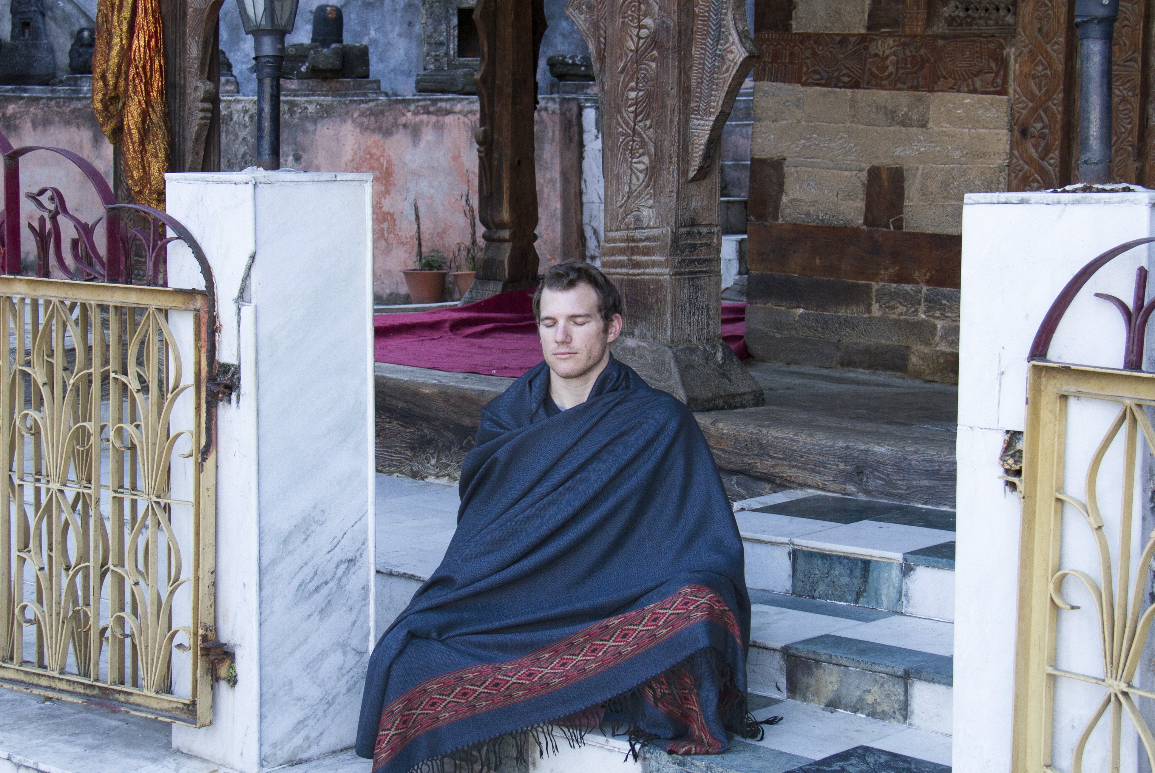 Person draped in a dark blue meditation shawl standing against a background of intricate temple woodwork carvings.