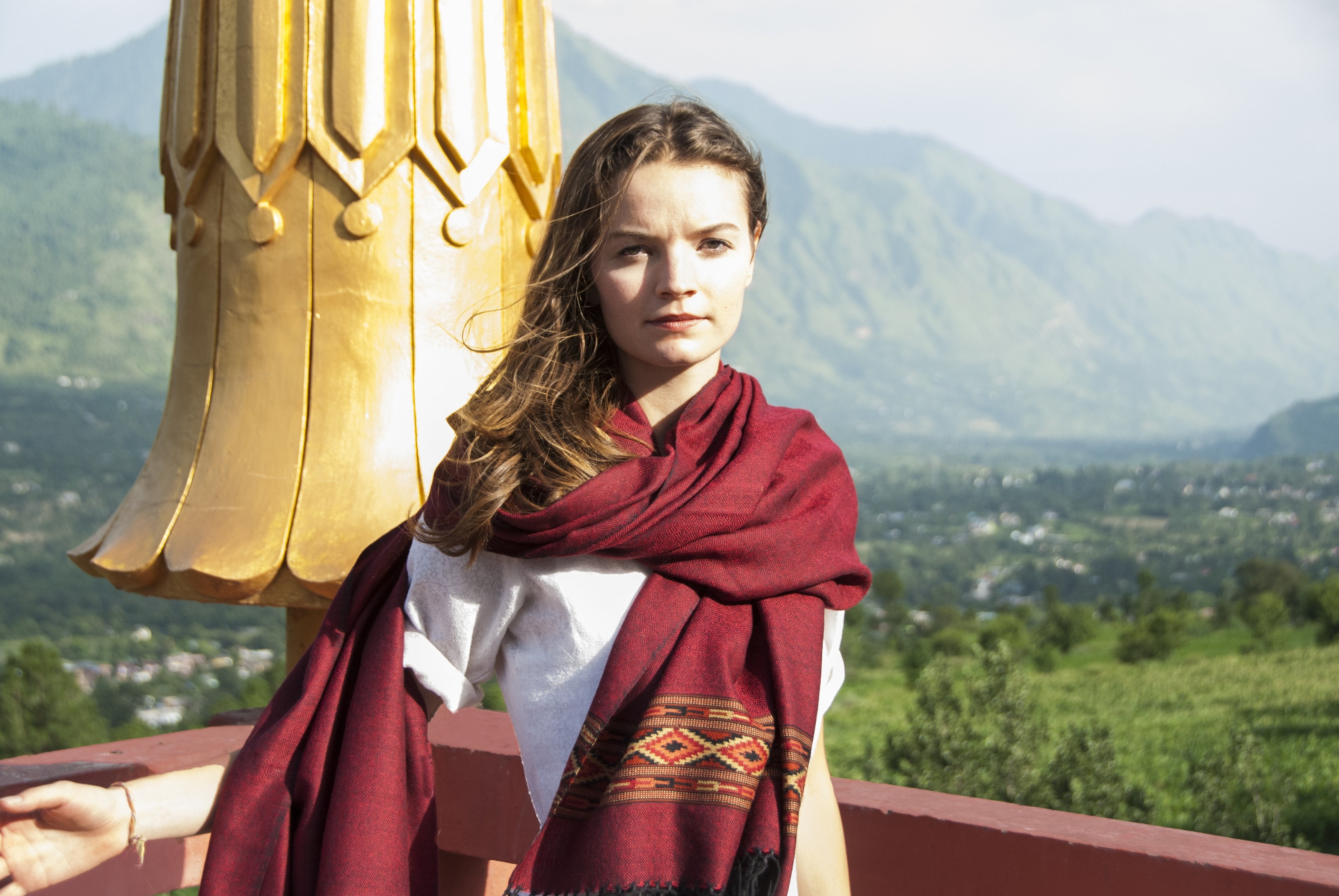 Individual with a dark red meditation shawl featuring ethnic patterns, posing against an ornately carved wooden temple wall.