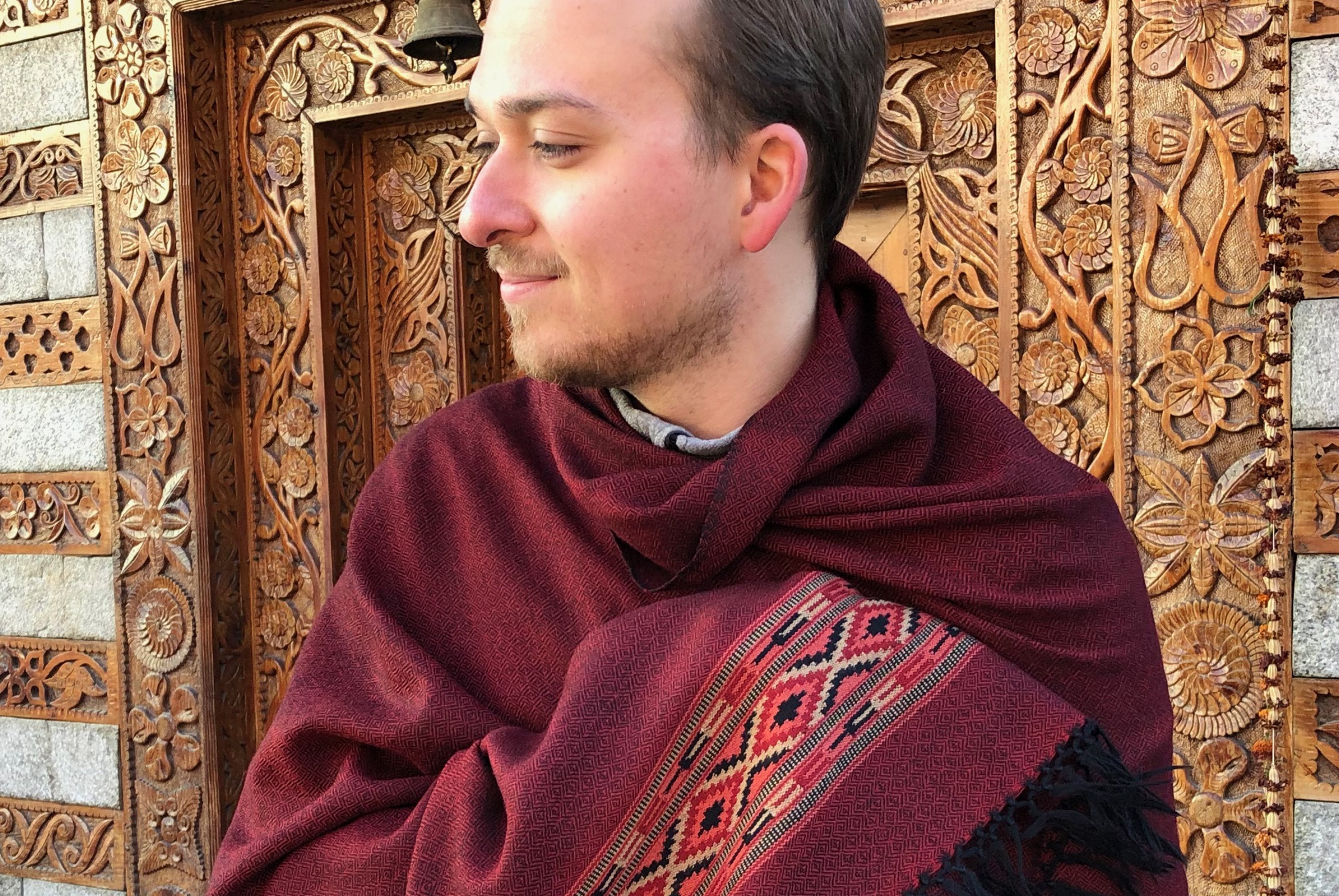 Individual with a dark red meditation shawl featuring ethnic patterns, posing against an ornately carved wooden temple wall.