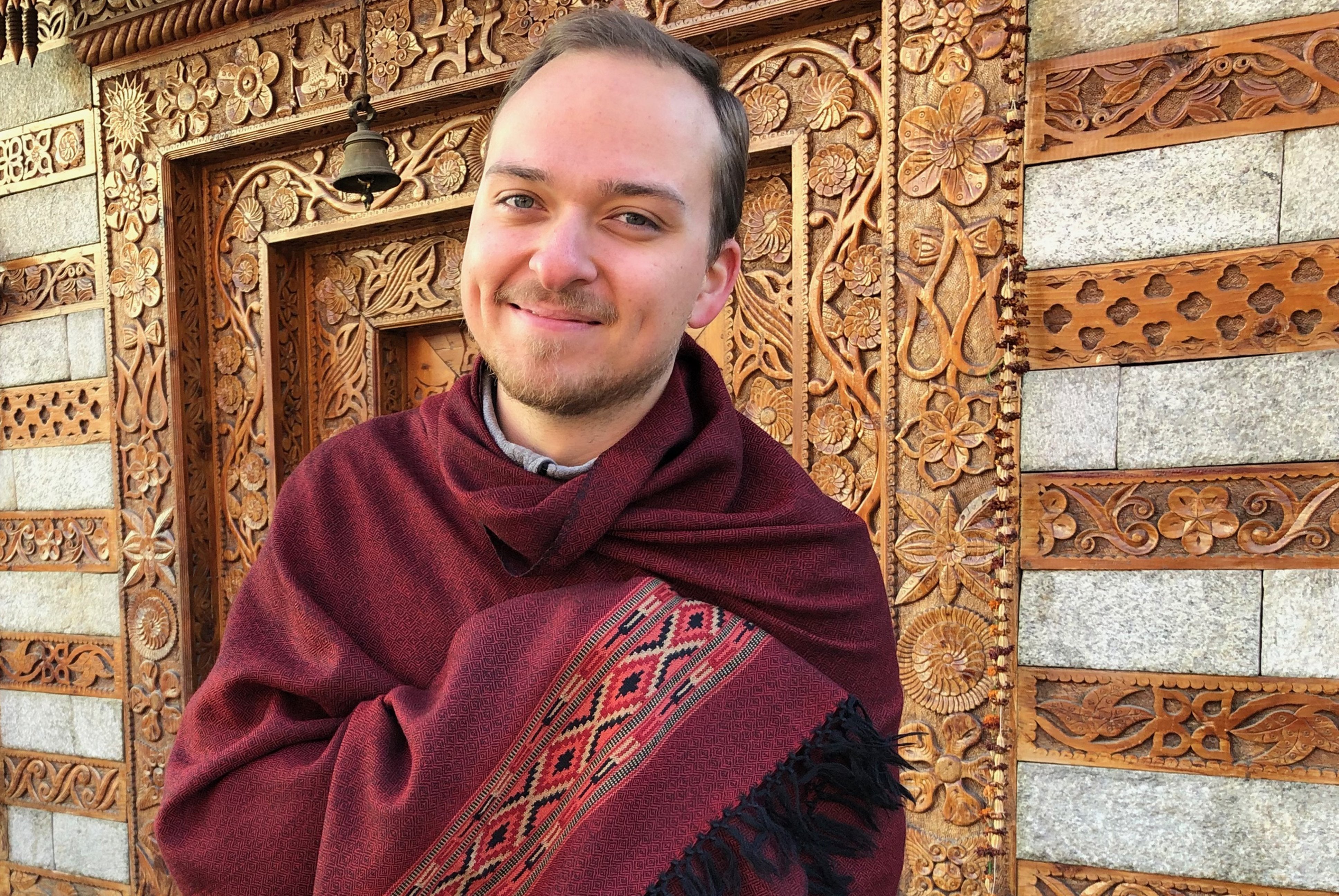 Person draped in a dark red meditation shawl standing against a background of intricate temple woodwork carvings.