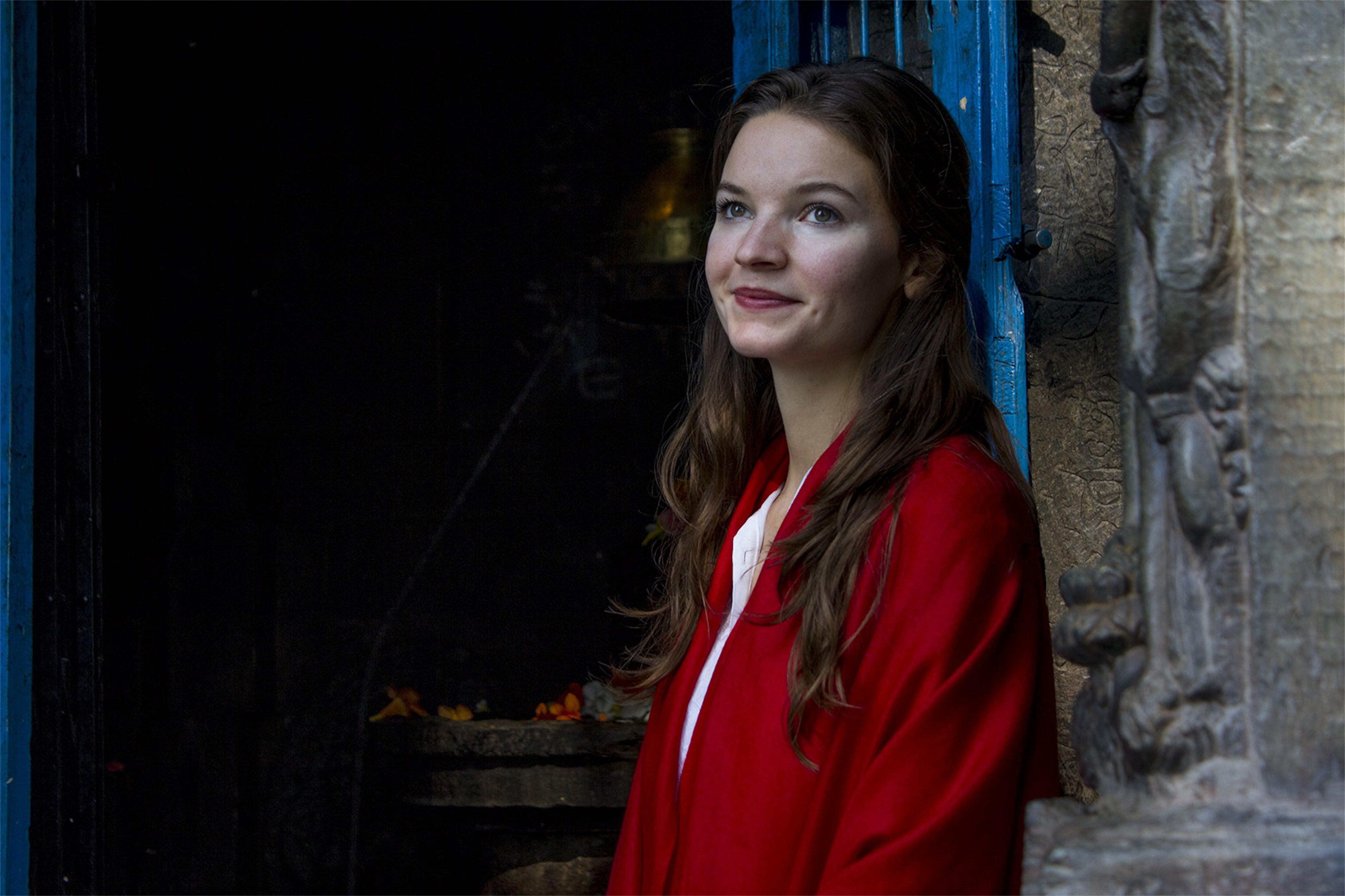 Young woman in a deep red pashmina meditation shawl, leaning against a blue temple door with a soft smile.