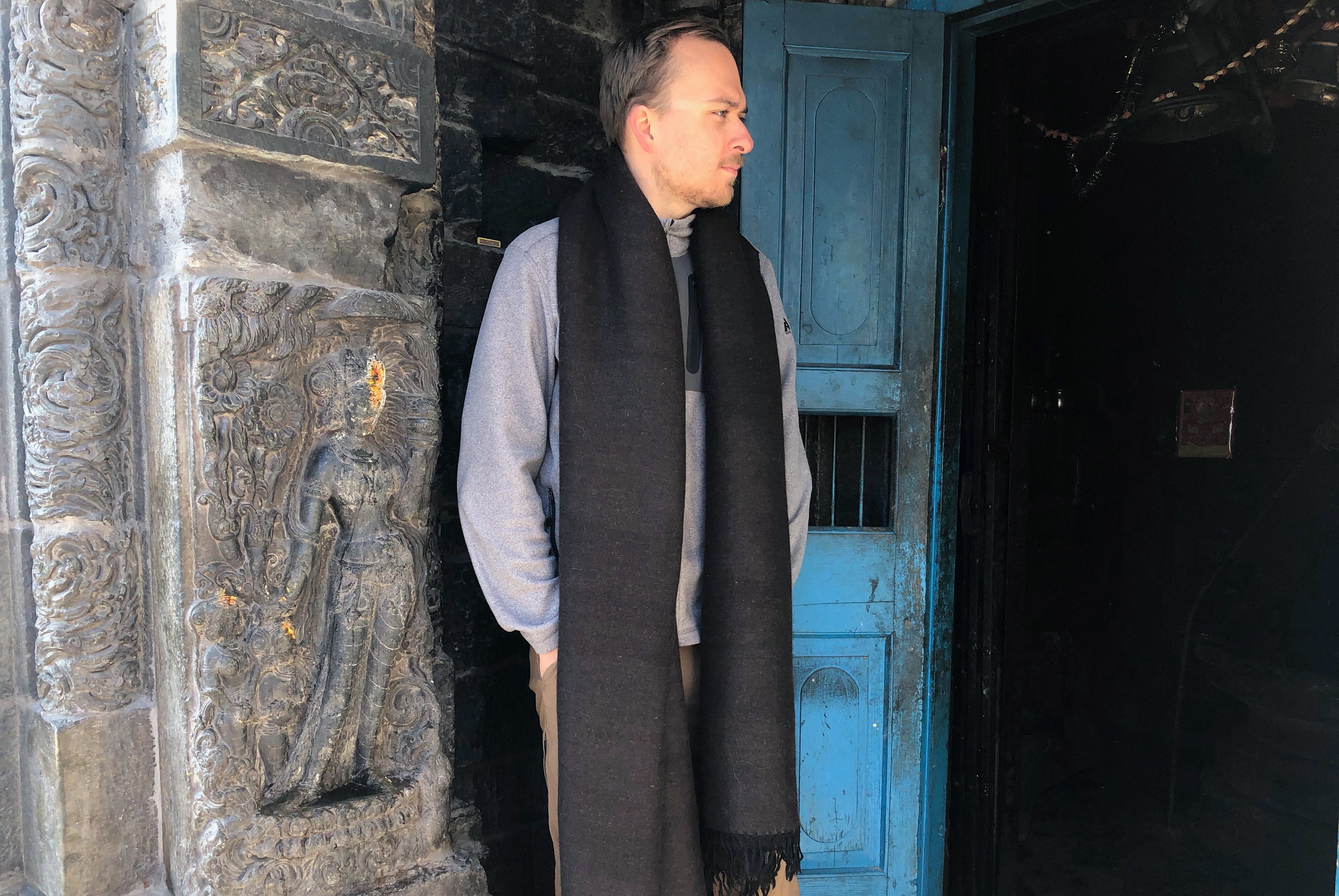 A man wearing a black Himalayan yak wool scarf, standing near the detailed stone carvings of a temple entrance with a blue wooden door.