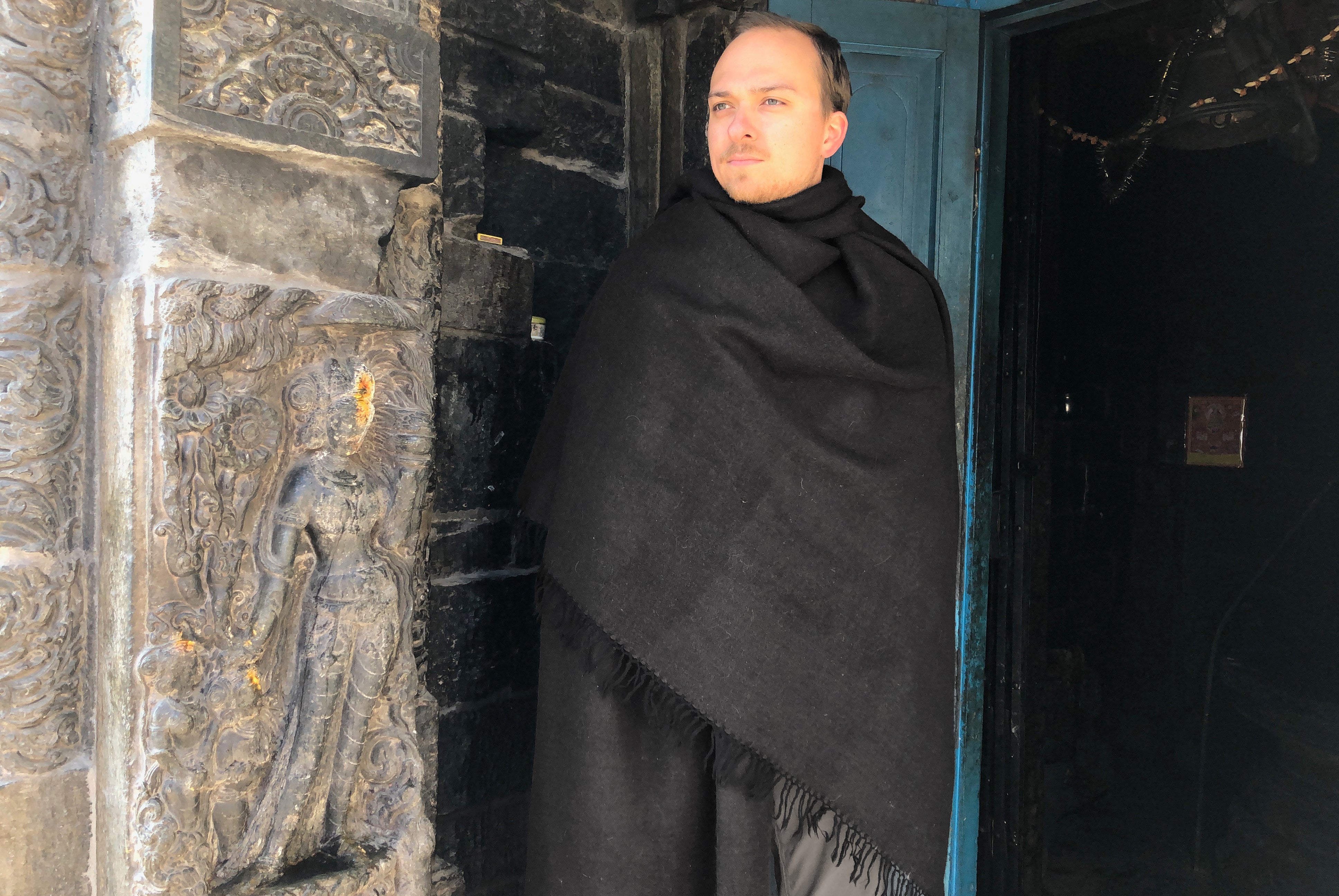 A man wrapped in a black Himalayan yak wool shawl, standing by the intricately carved stone entrance of a temple.