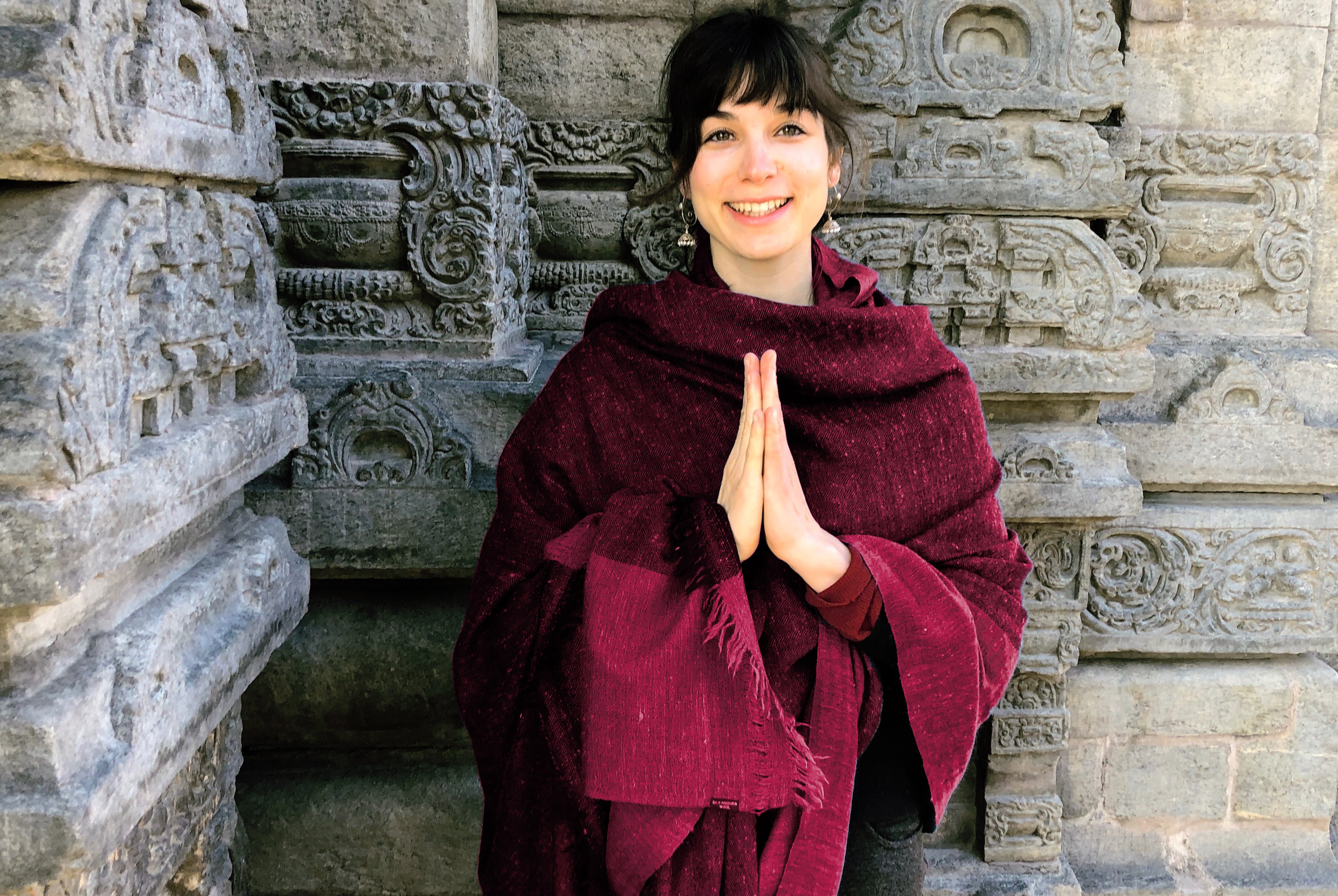 A person wearing a handwoven Ishwar meditation blanket in rich burgundy color, standing by intricately carved stonework, hands in a prayer gesture.