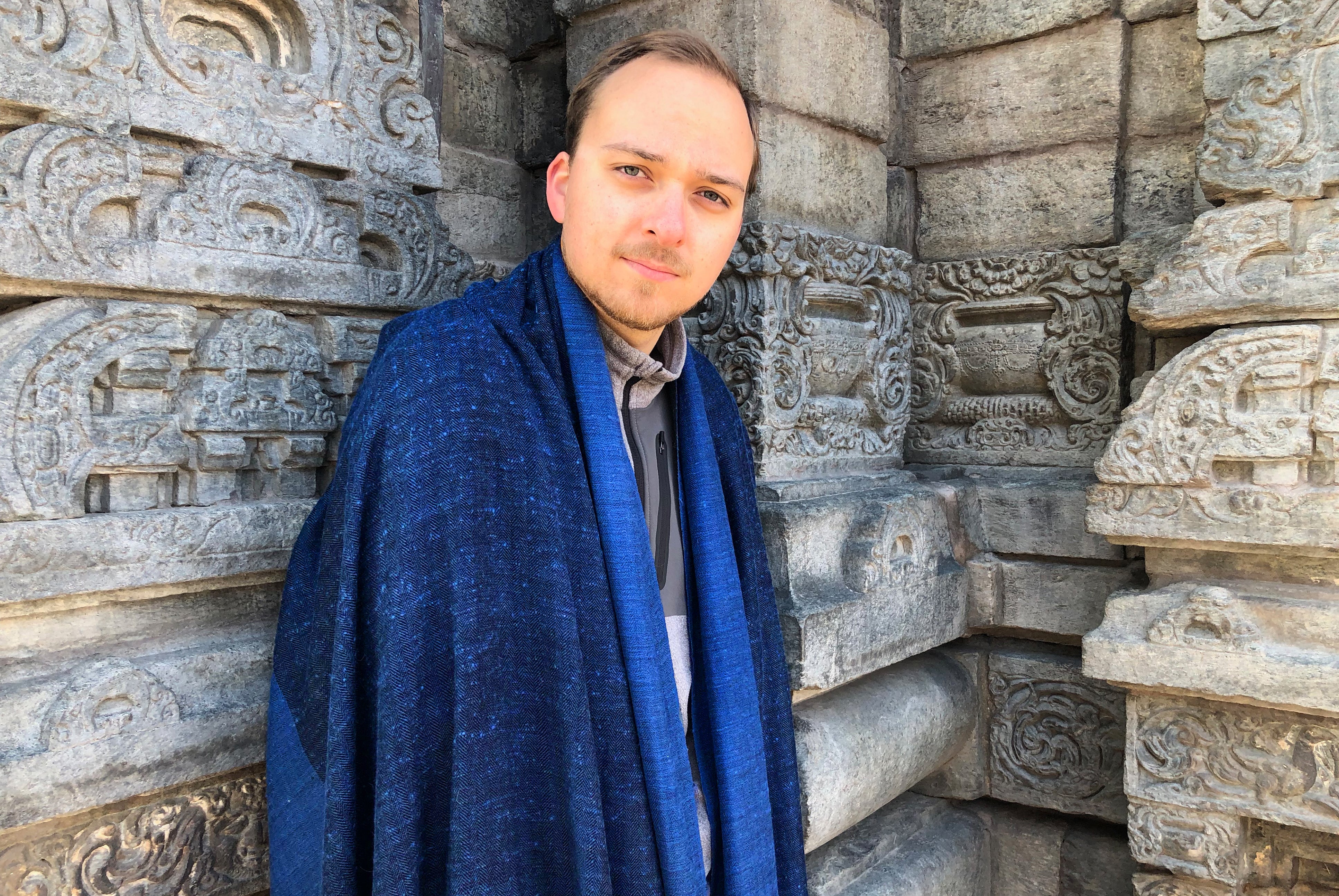 Man wearing a blue shawl, standing against a stone background with a calm expression.