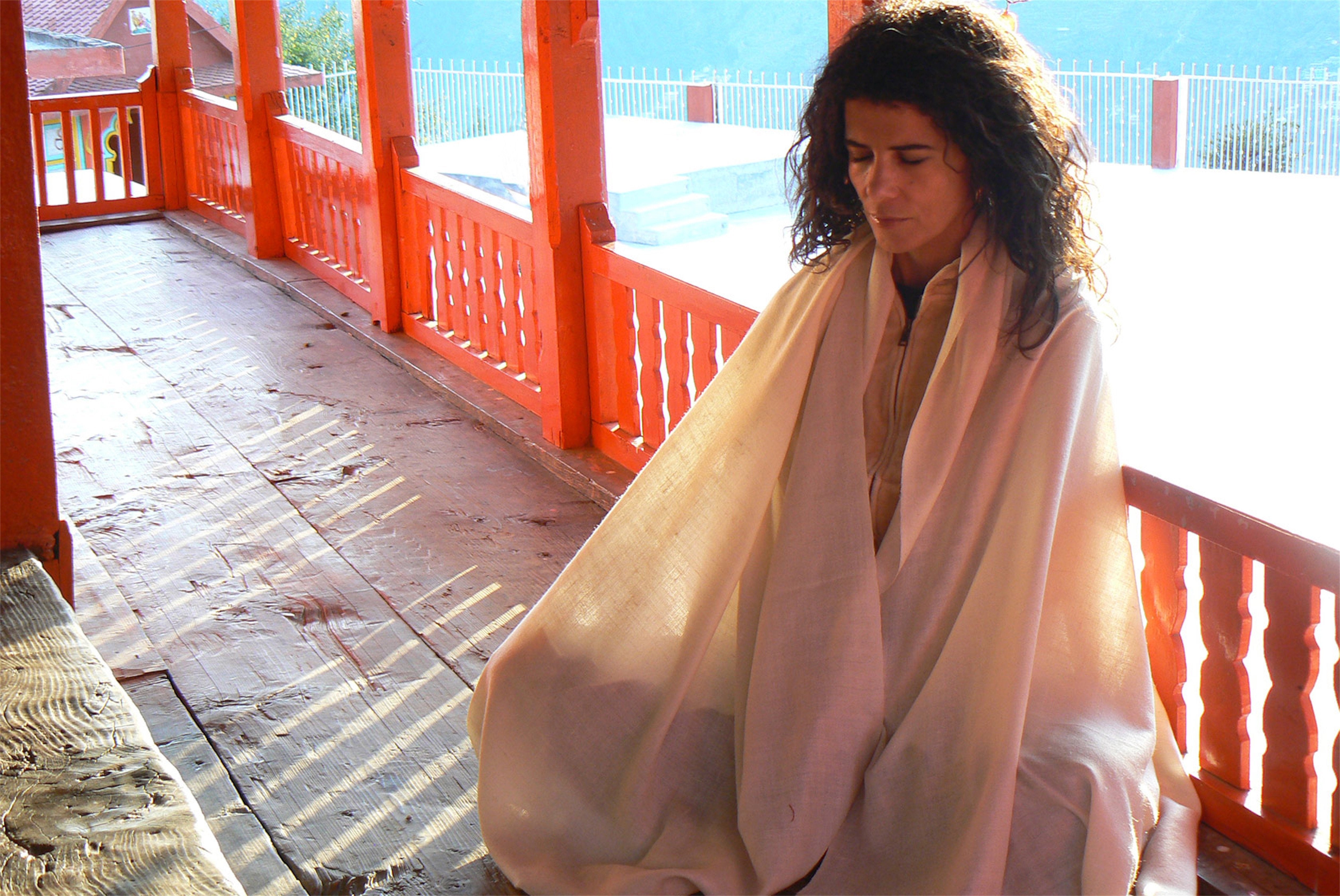 Woman meditating outdoors in a cream shawl, seated on a porch with an orange railing and sunlight streaming through.