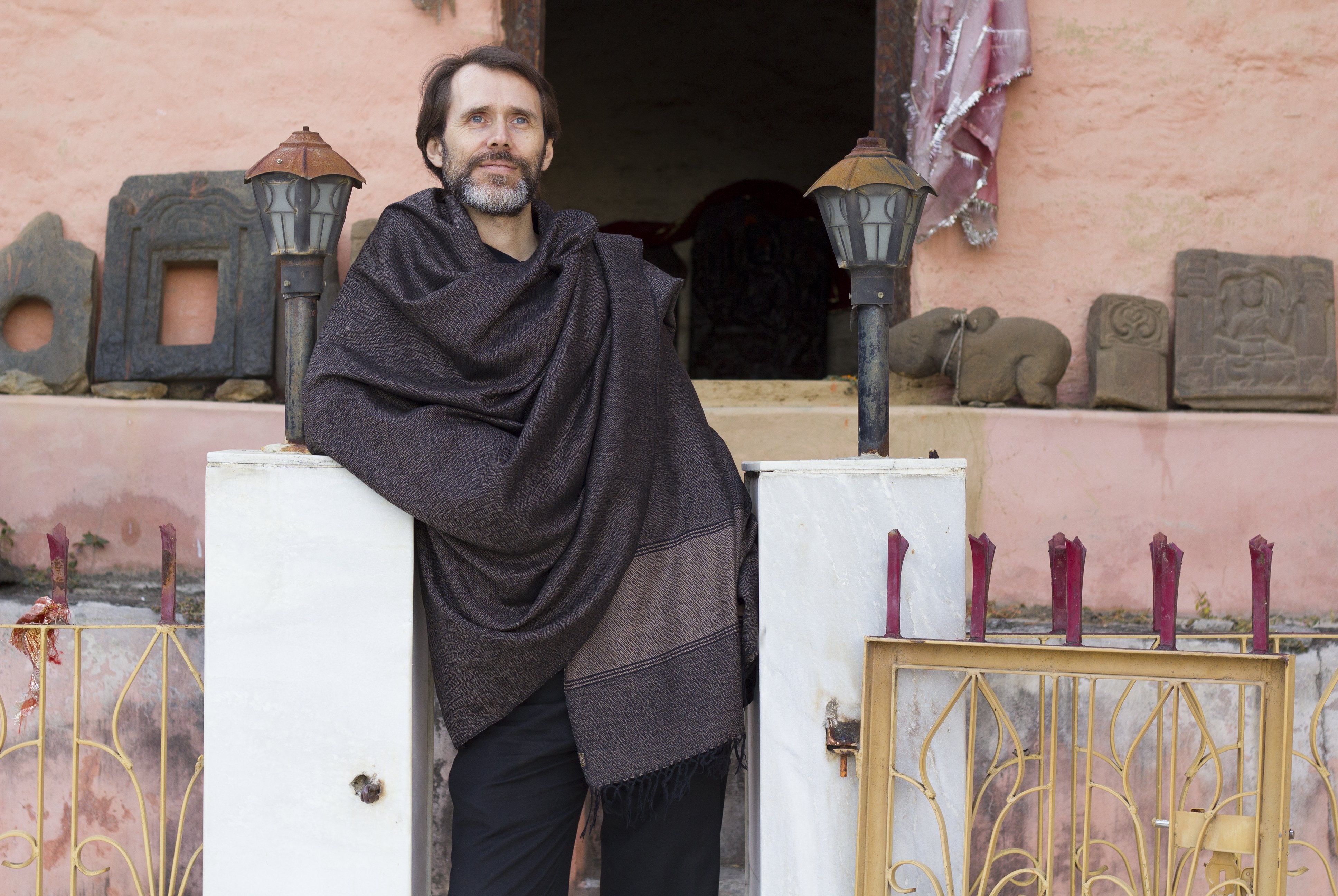 A man standing calmly in a rustic setting, wrapped in a dark brown Milarepa large meditation shawl, with a backdrop of ancient stone carvings and soft lighting.