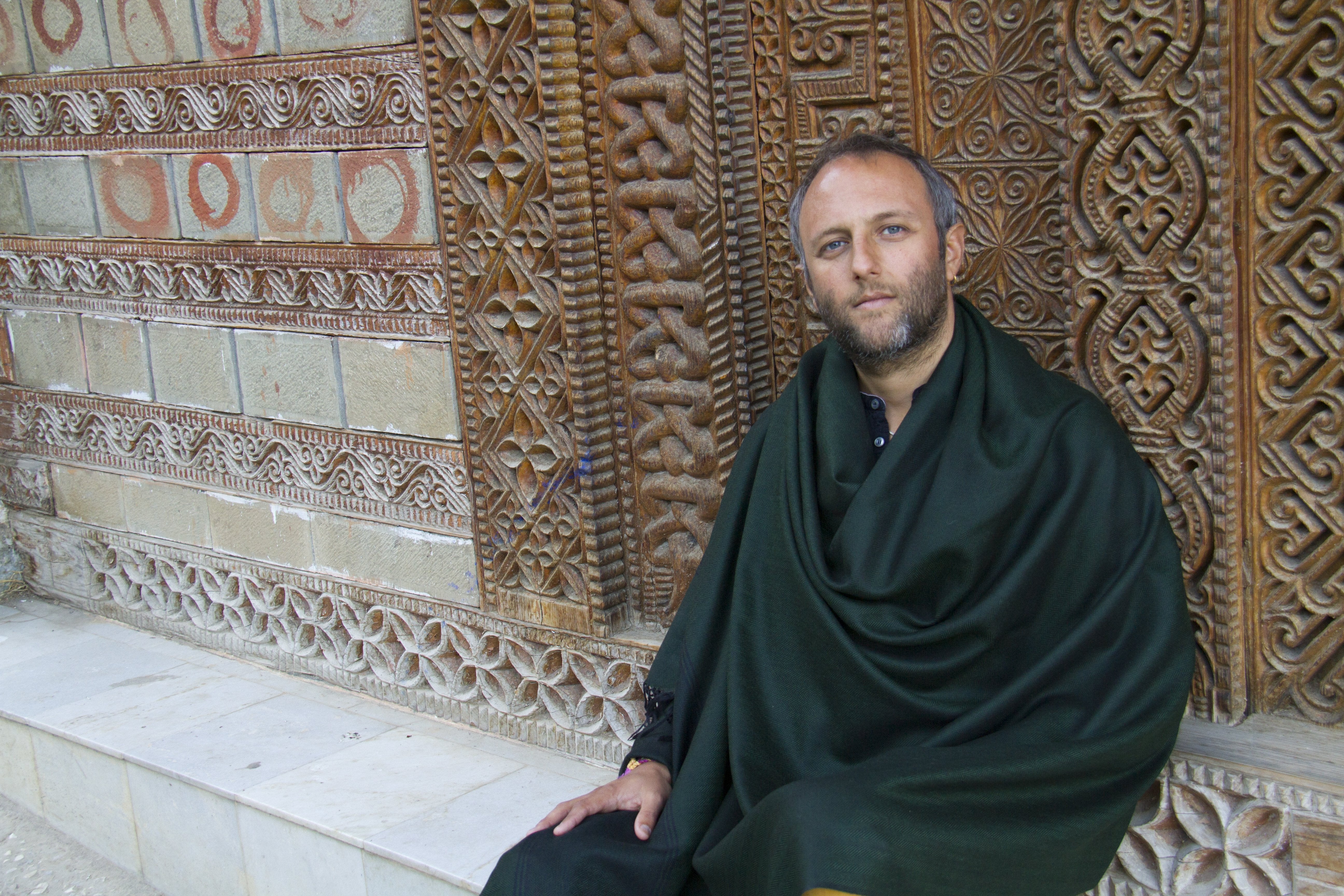 A man wrapped in a deep green Milarepa wool meditation shawl, seated against an intricately carved wooden wall with traditional patterns.