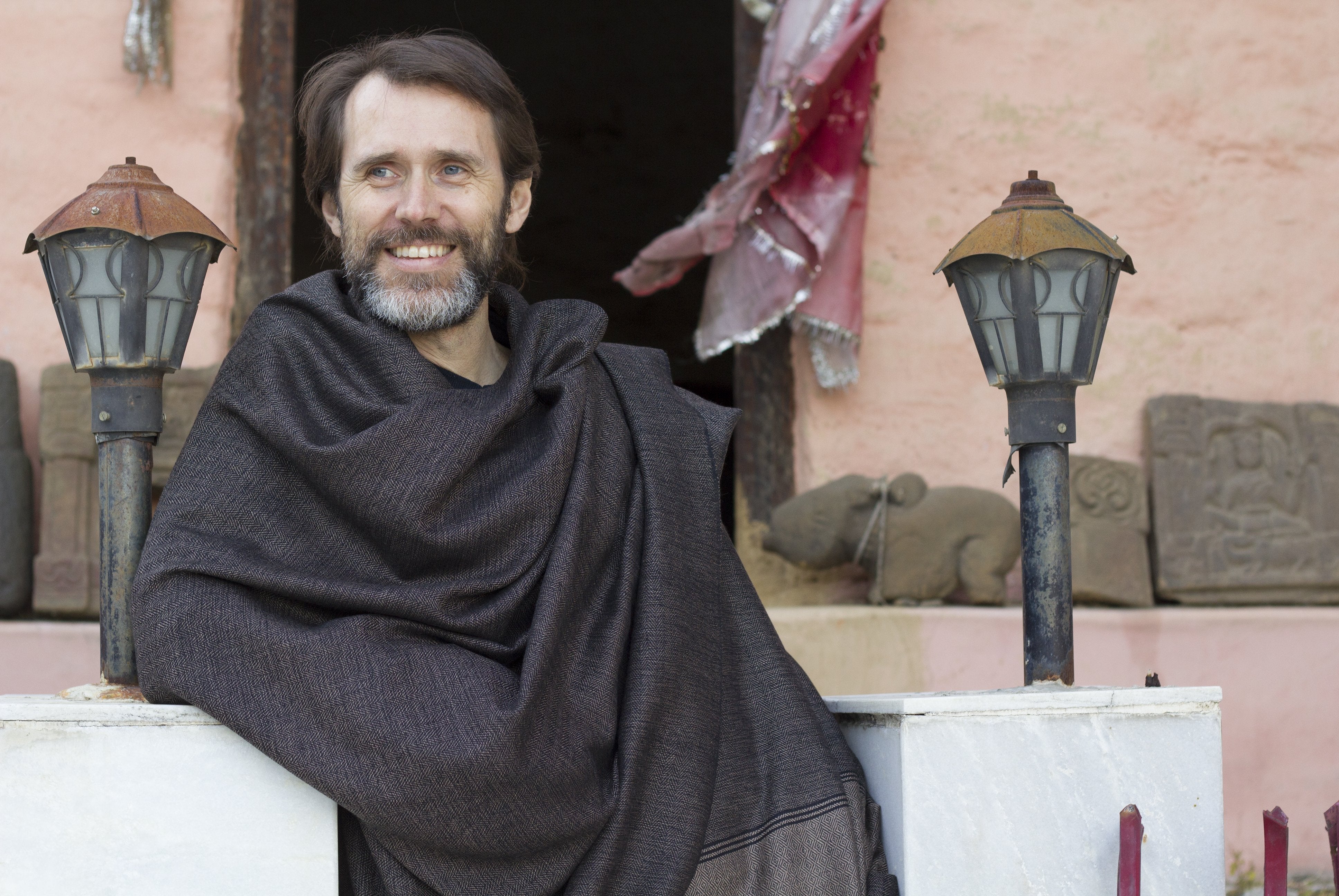 A man smiling while draped in a dark brown Milarepa large wool shawl, standing next to rustic lanterns and ancient stone carvings, exuding a serene and contemplative vibe.