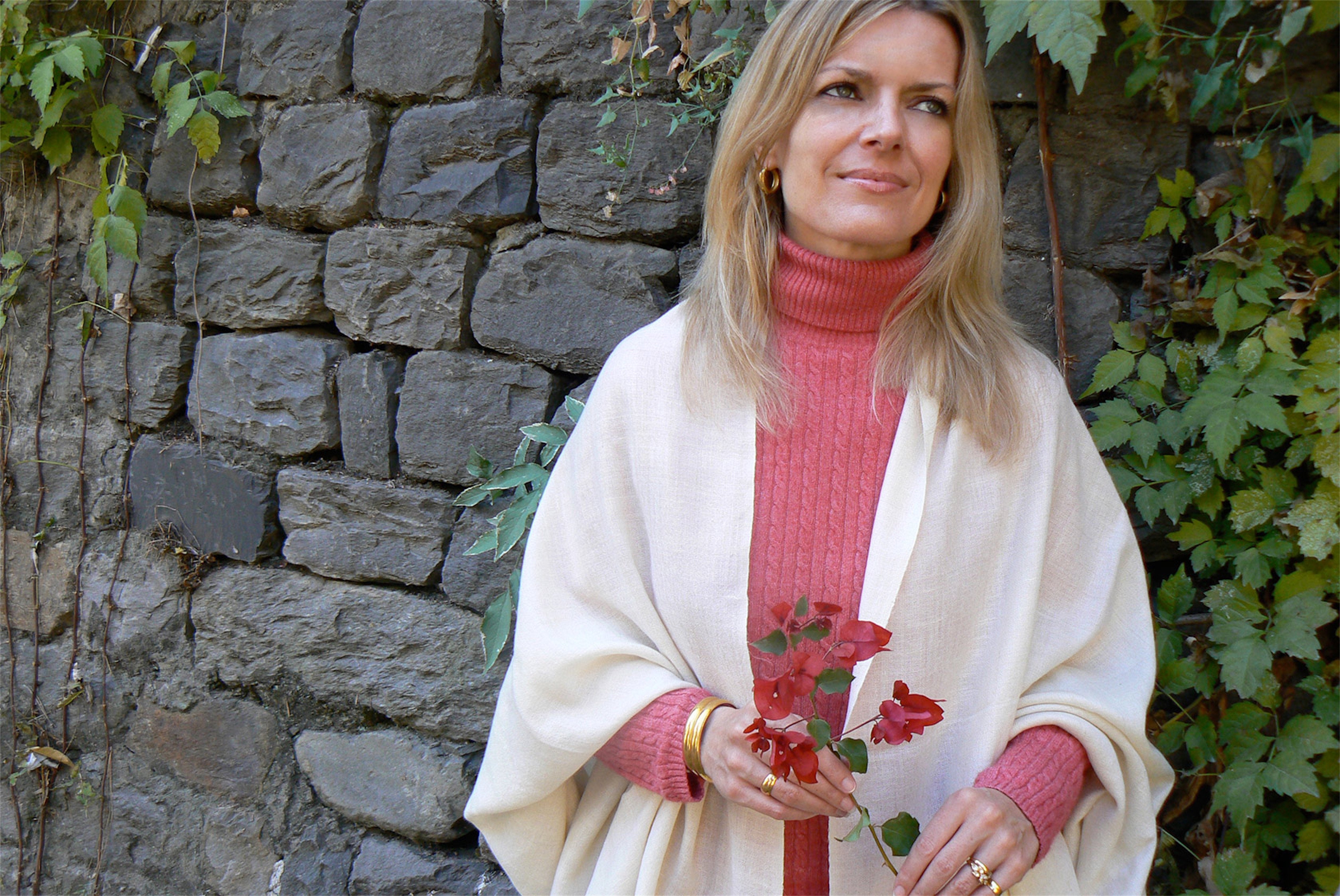 Portrait of a woman in a cream shawl with a red sweater, standing by a stone wall and holding red flowers.