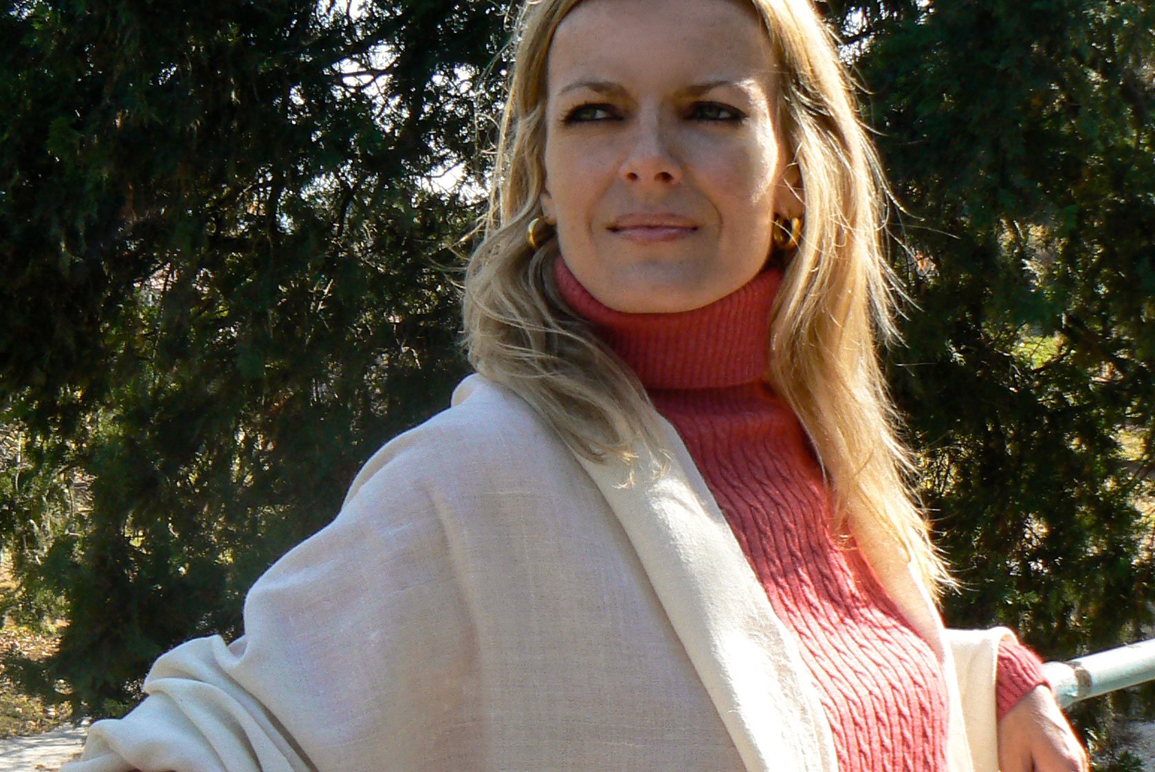 Woman in a cream shawl with a red sweater, gazing into the distance with greenery in the background.