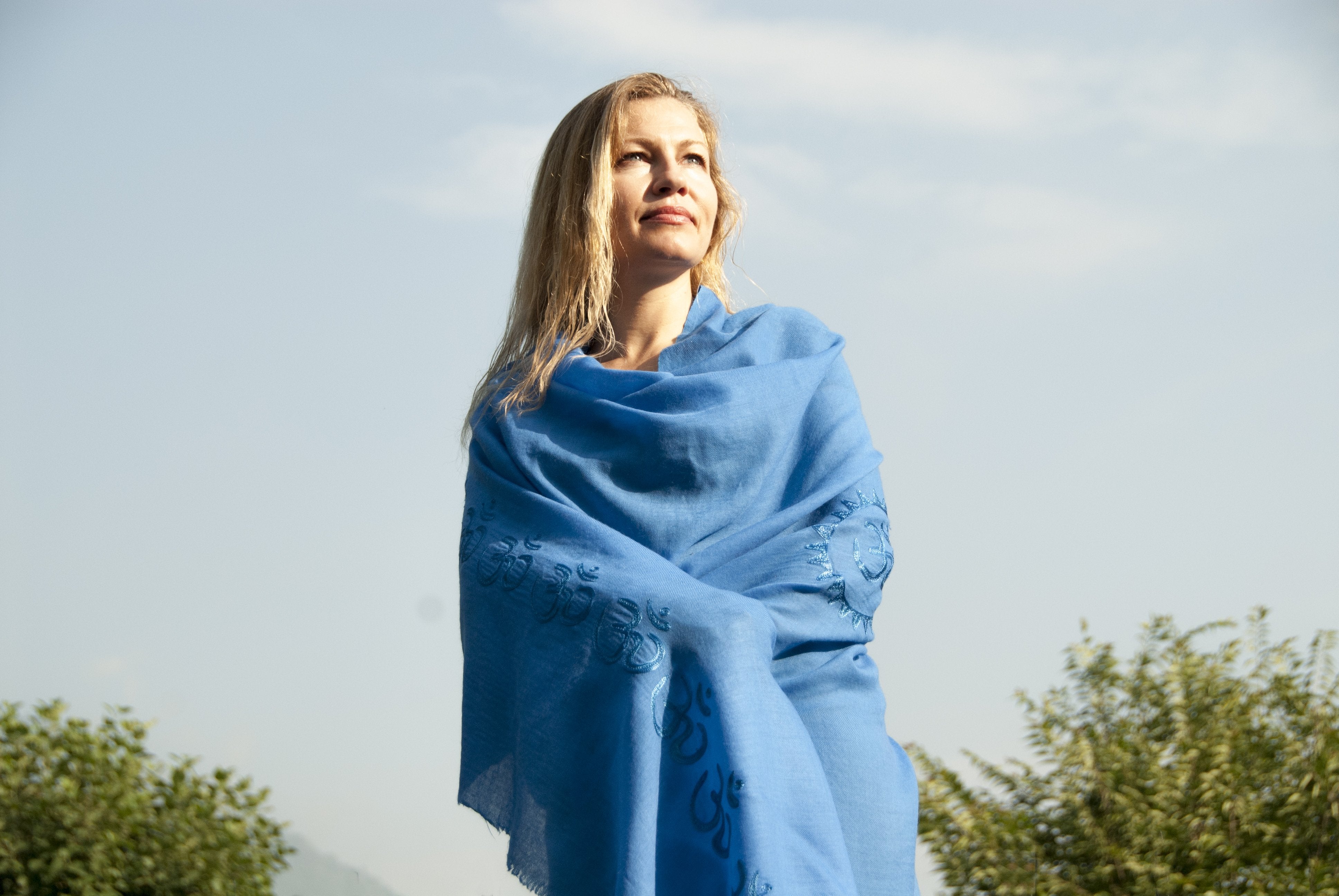 Woman standing outdoors wrapped in a serene blue Padma shawl, adorned with intricately embroidered Om symbols, gazing upwards against a clear blue sky with lush green trees in the background.