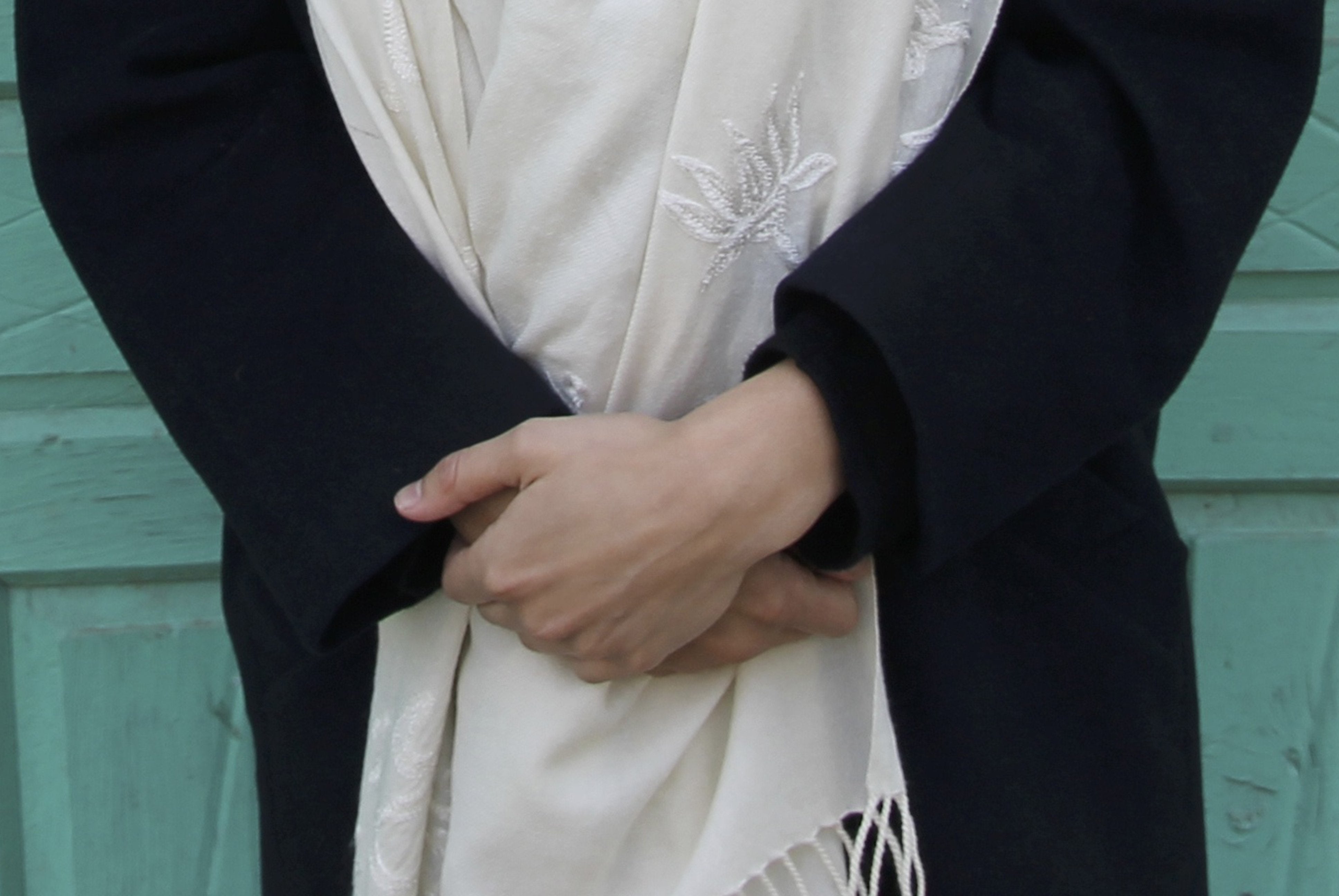 Close-up of hands gently holding a soft white Padma meditation shawl, adorned with elegant embroidered lotus designs, draped over a dark coat against a teal background.