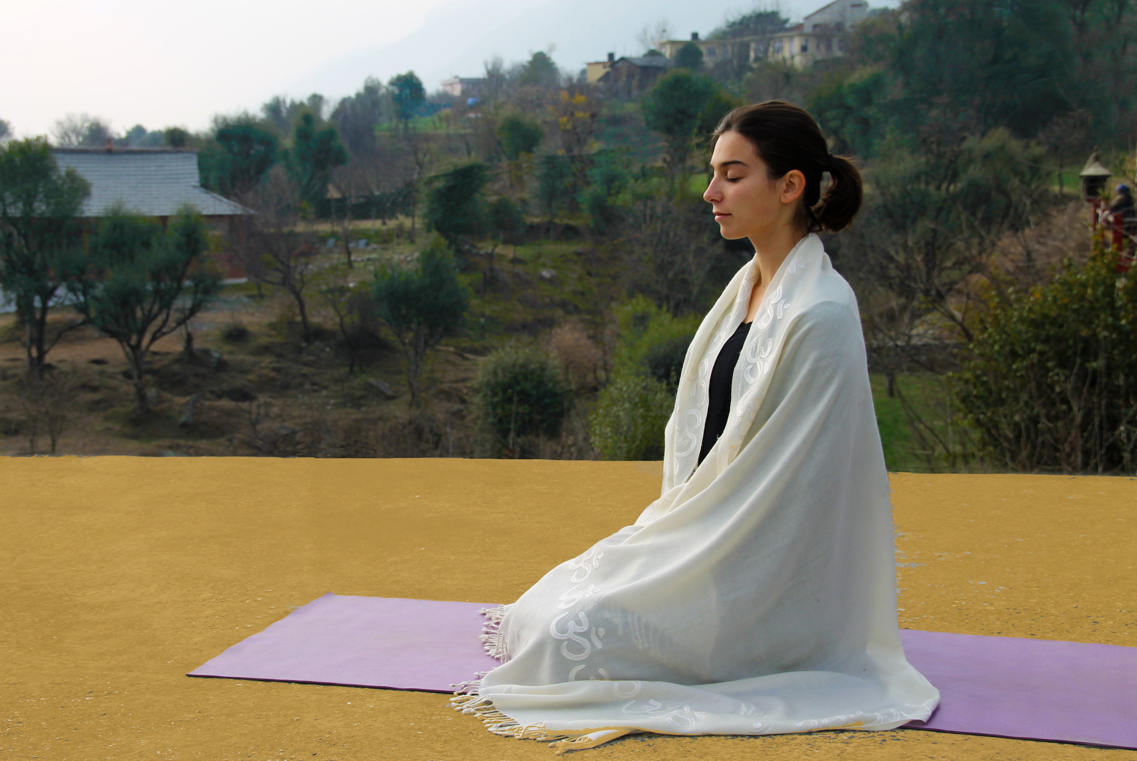 Woman meditating outdoors with a white Padma shawl adorned with Om symbols draped over her shoulders, seated on a purple yoga mat against a serene natural backdrop of trees and distant houses.