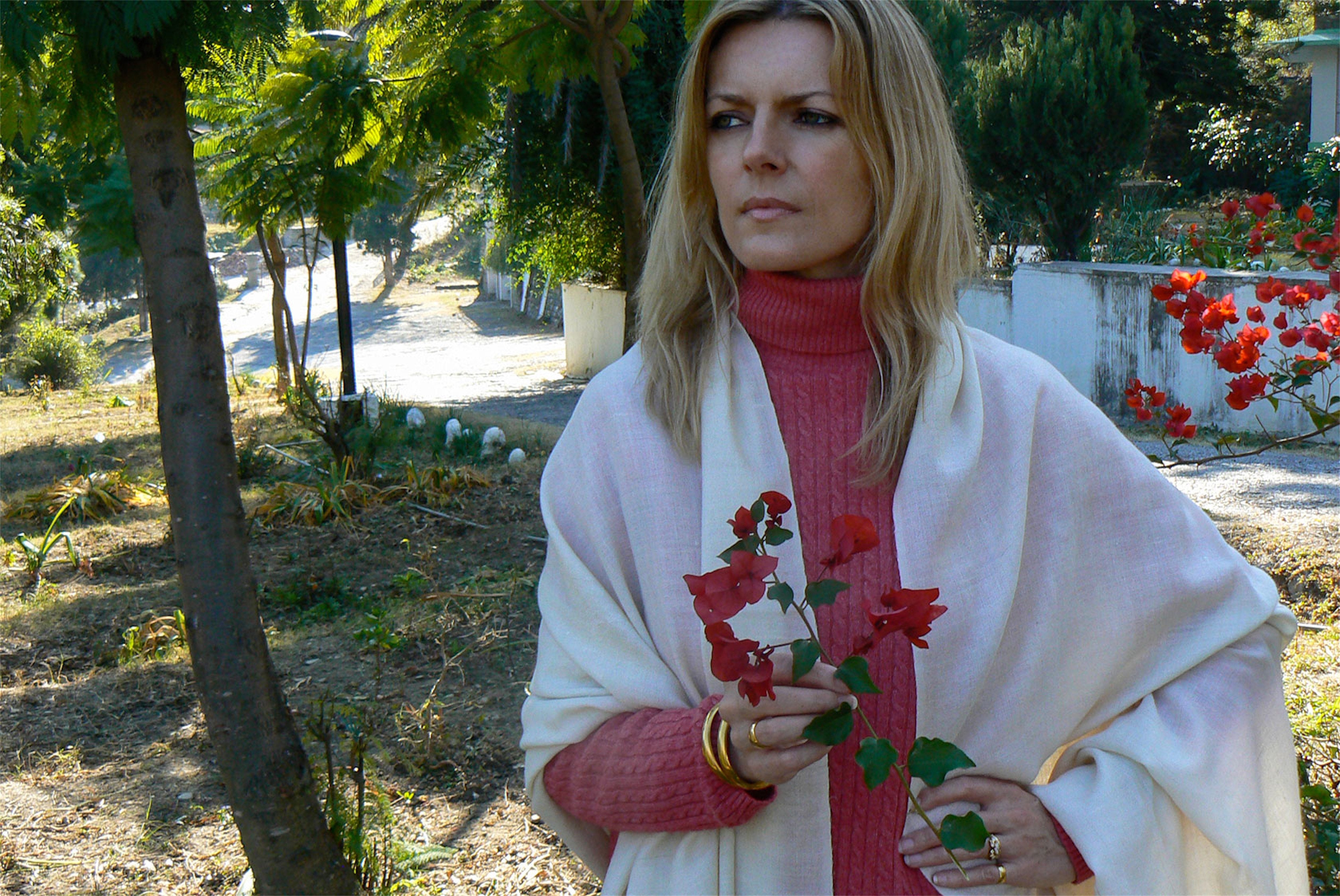 Woman with a cream pashmina meditation shawl in a garden, holding bougainvillea flowers with a pensive expression.