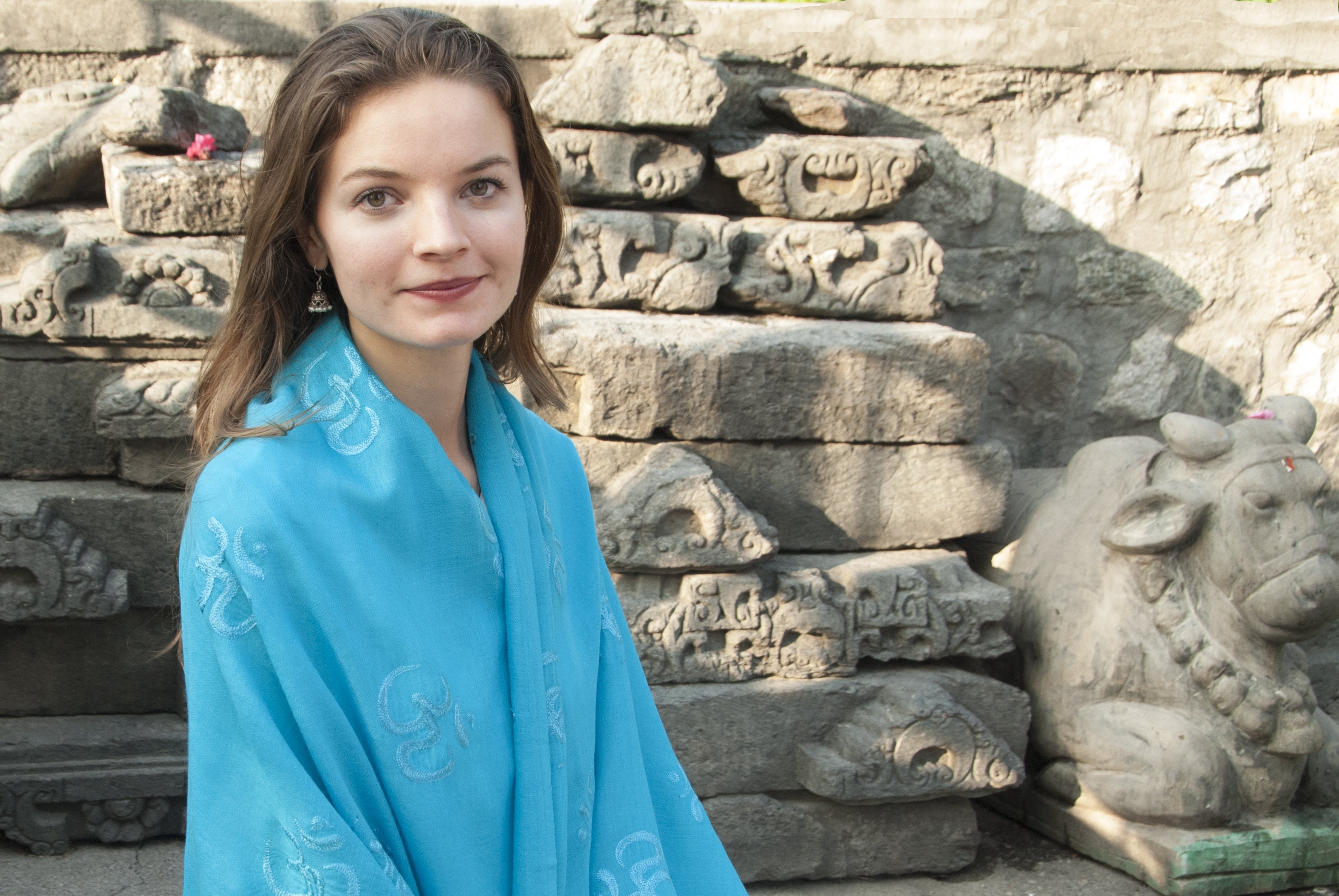 Woman seated gracefully, draped in a vibrant blue Purna yoga shawl with intricate Om symbols embroidered, set against a backdrop of ancient stone carvings and statues.