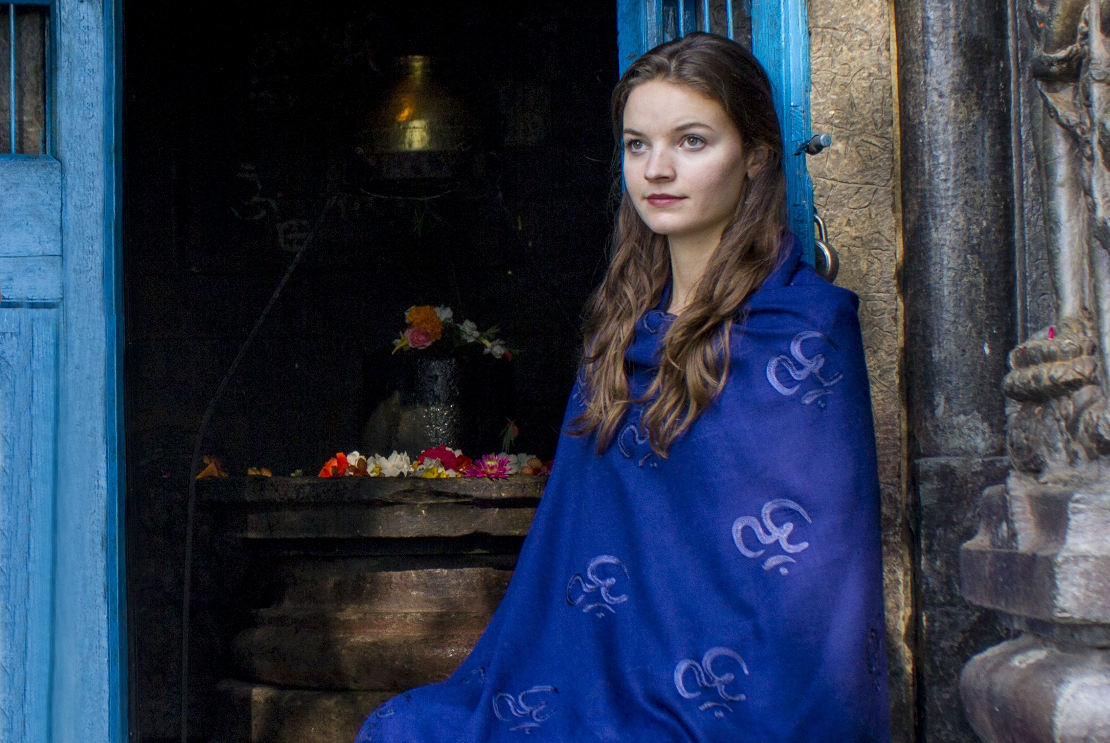 Woman seated by an intricately carved temple doorway, wrapped in a vibrant blue Purna yoga shawl adorned with Om embroidery, with floral offerings visible in the background.