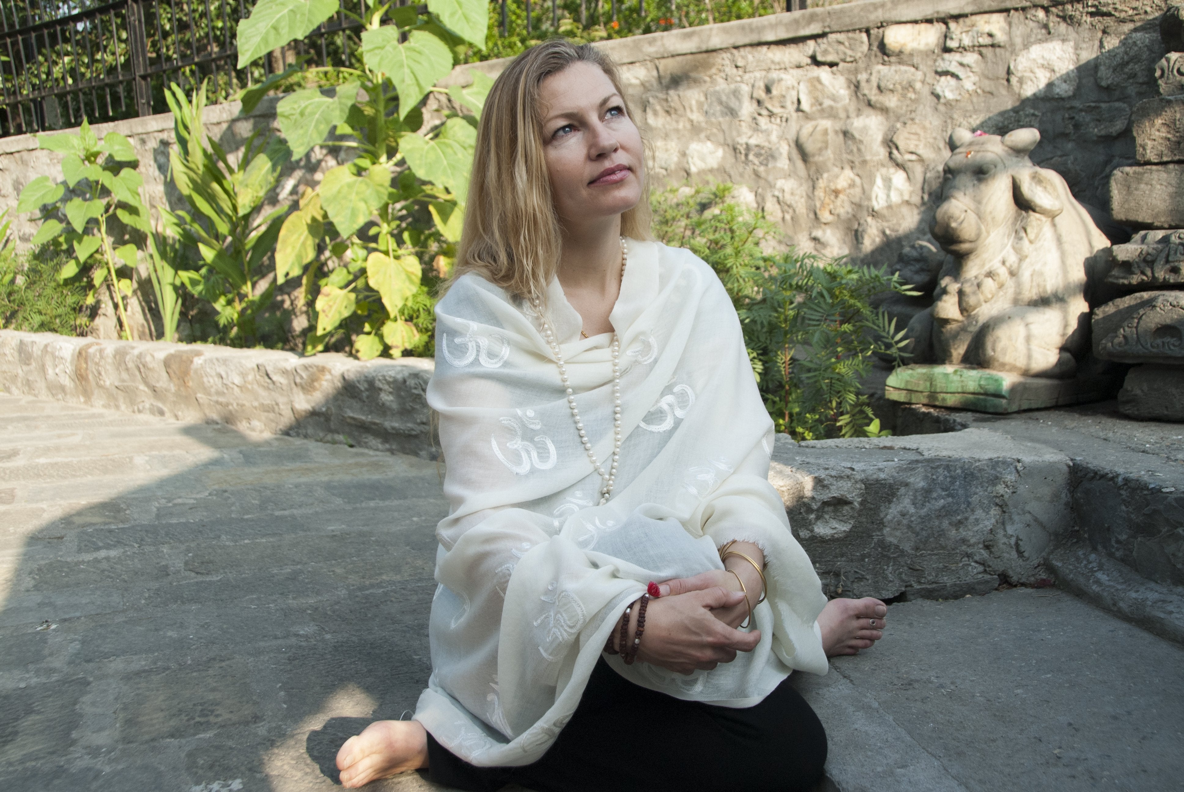 Woman seated on stone steps, wrapped in a serene white Purna yoga shawl with delicate Om symbols embroidered, meditating peacefully outdoors beside ancient stone carvings and lush greenery.