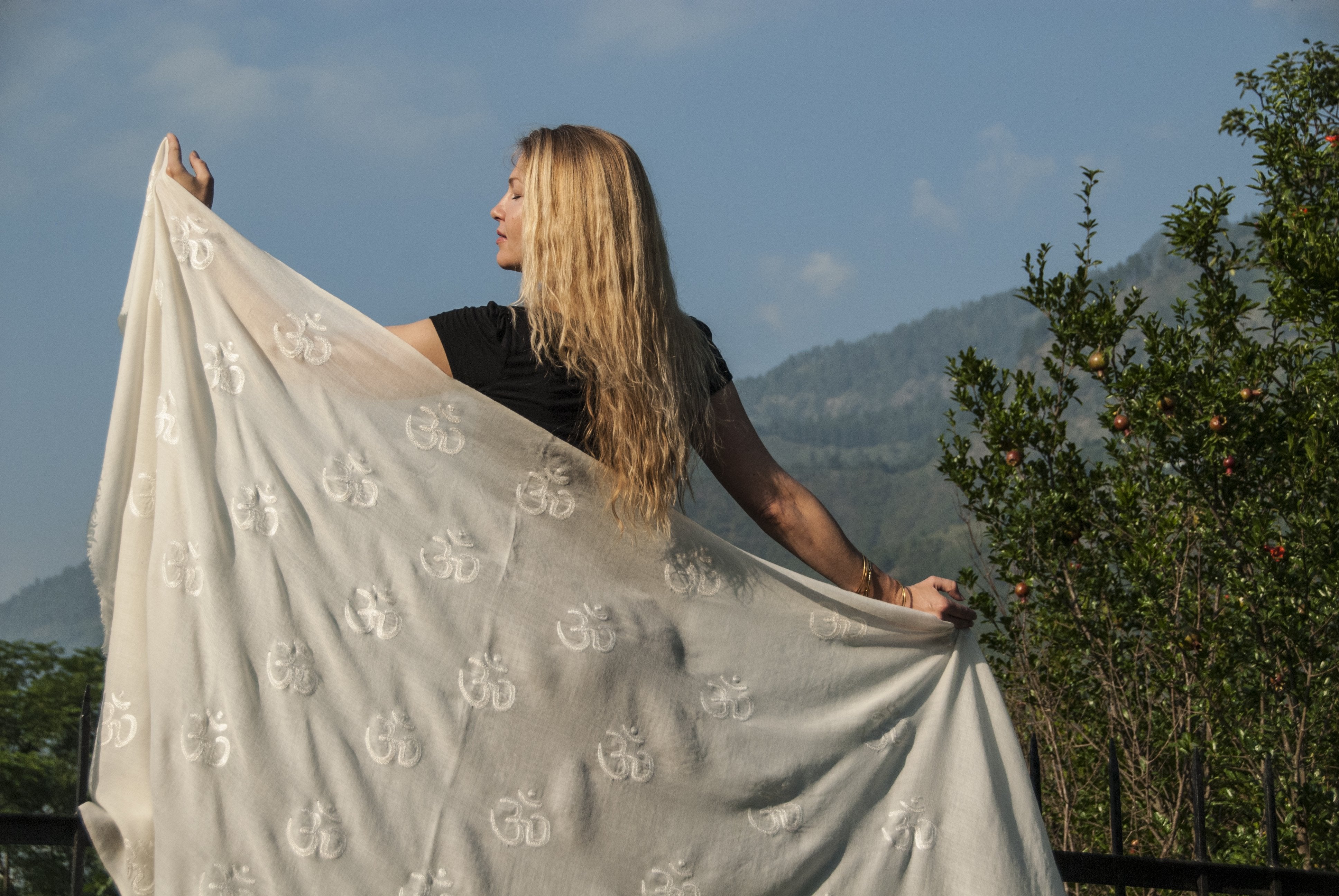 Woman gracefully holding a flowing white Purna yoga shawl with Om embroidery, standing outdoors against a serene mountain backdrop and lush greenery under a clear blue sky.