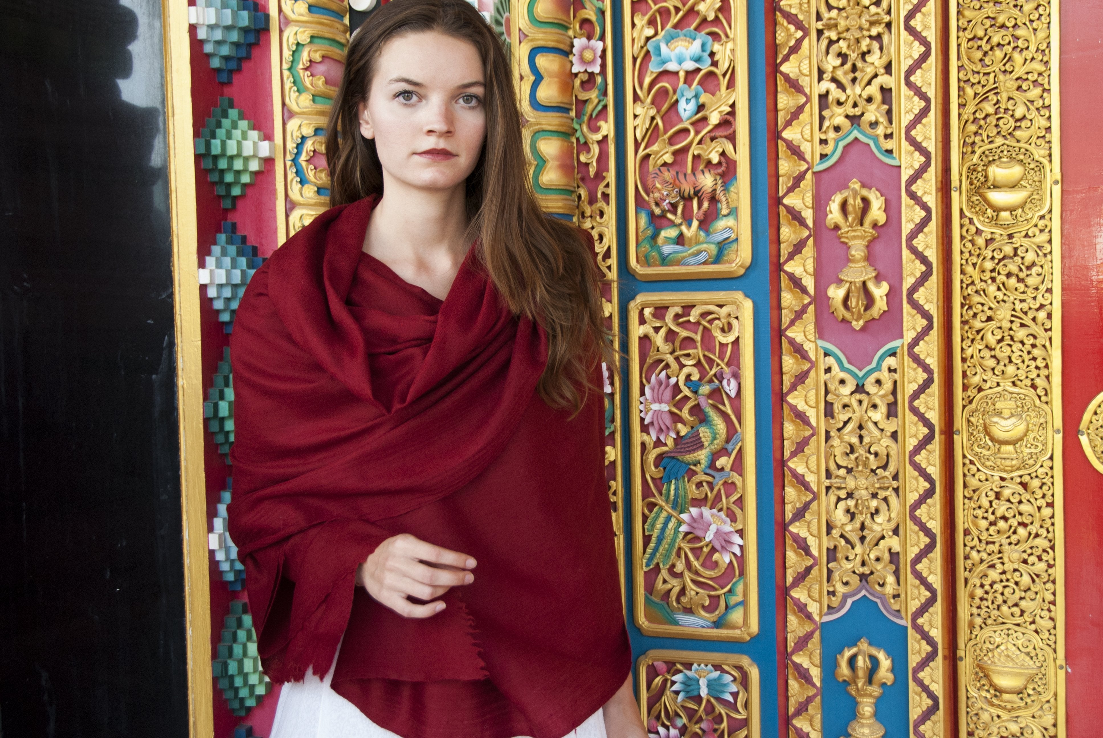 A young woman stands against a vibrant and intricately decorated temple wall, wearing a luxurious burgundy Rasana prayer shawl, crafted from soft angora wool, exuding elegance and tradition.