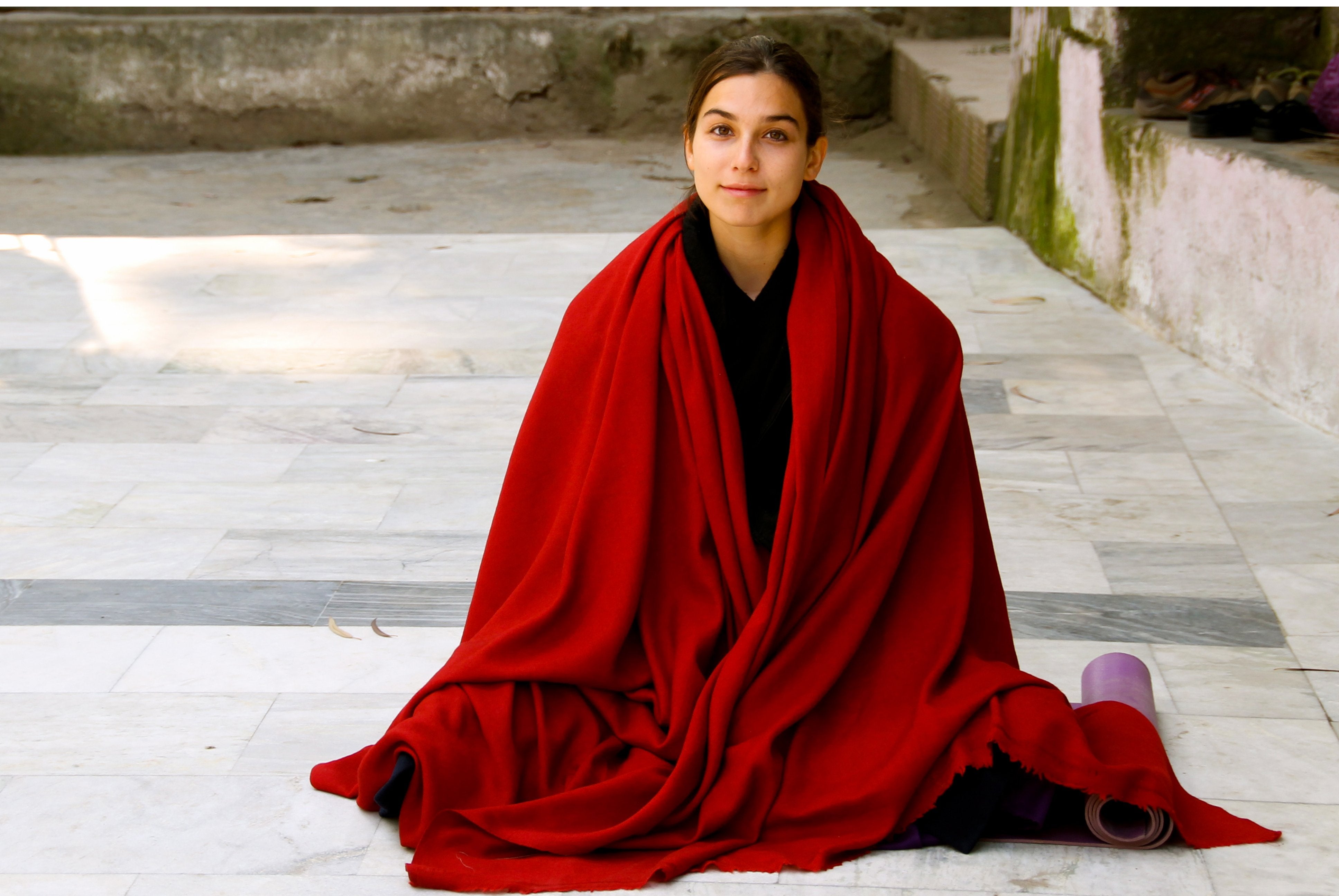 Young woman seated on a marble floor, draped in a bright red Rasana prayer shawl made of soft angora wool, with a yoga mat beside her, radiating serenity in a tranquil courtyard setting.