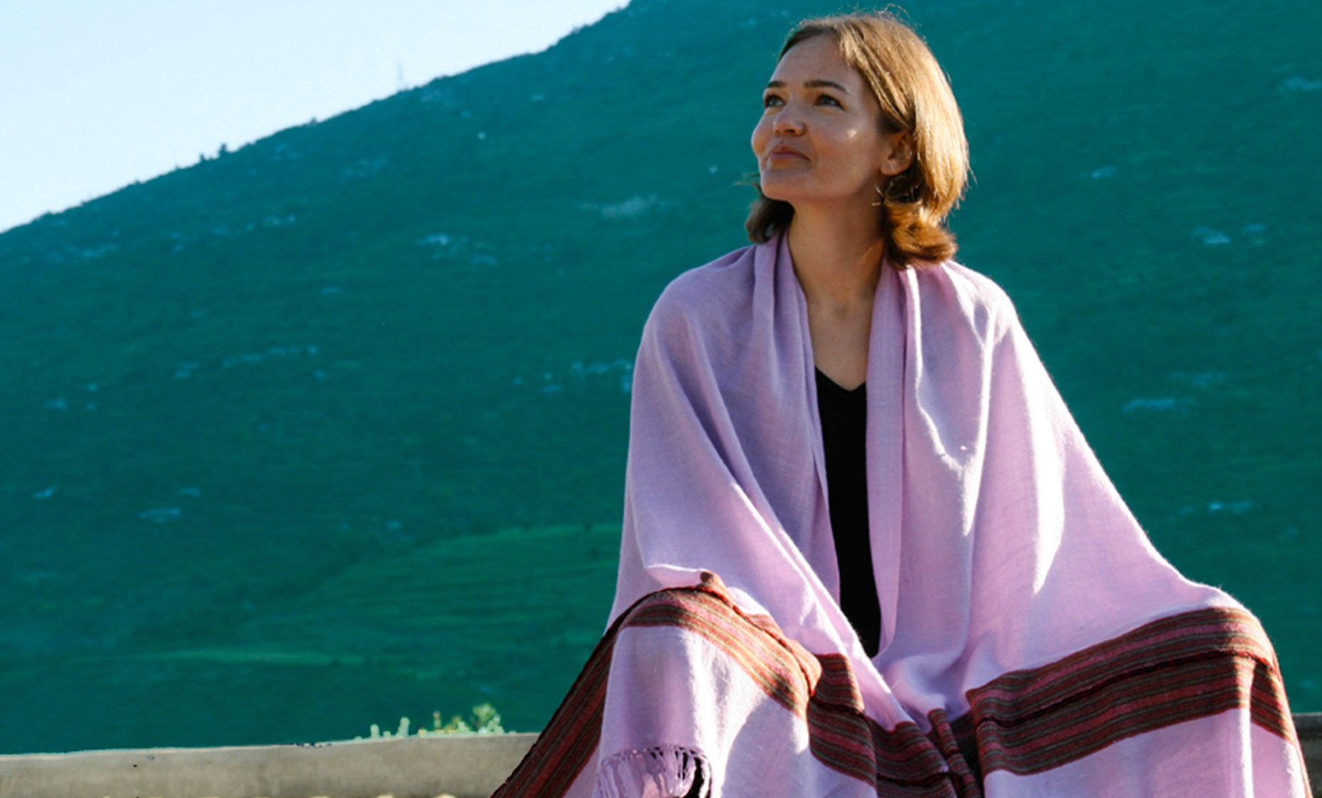 A tranquil image of a person adorned in a pink Santosh ethnic Indian meditation shawl. The backdrop of lush green mountains adds a peaceful and serene vibe to the composition.