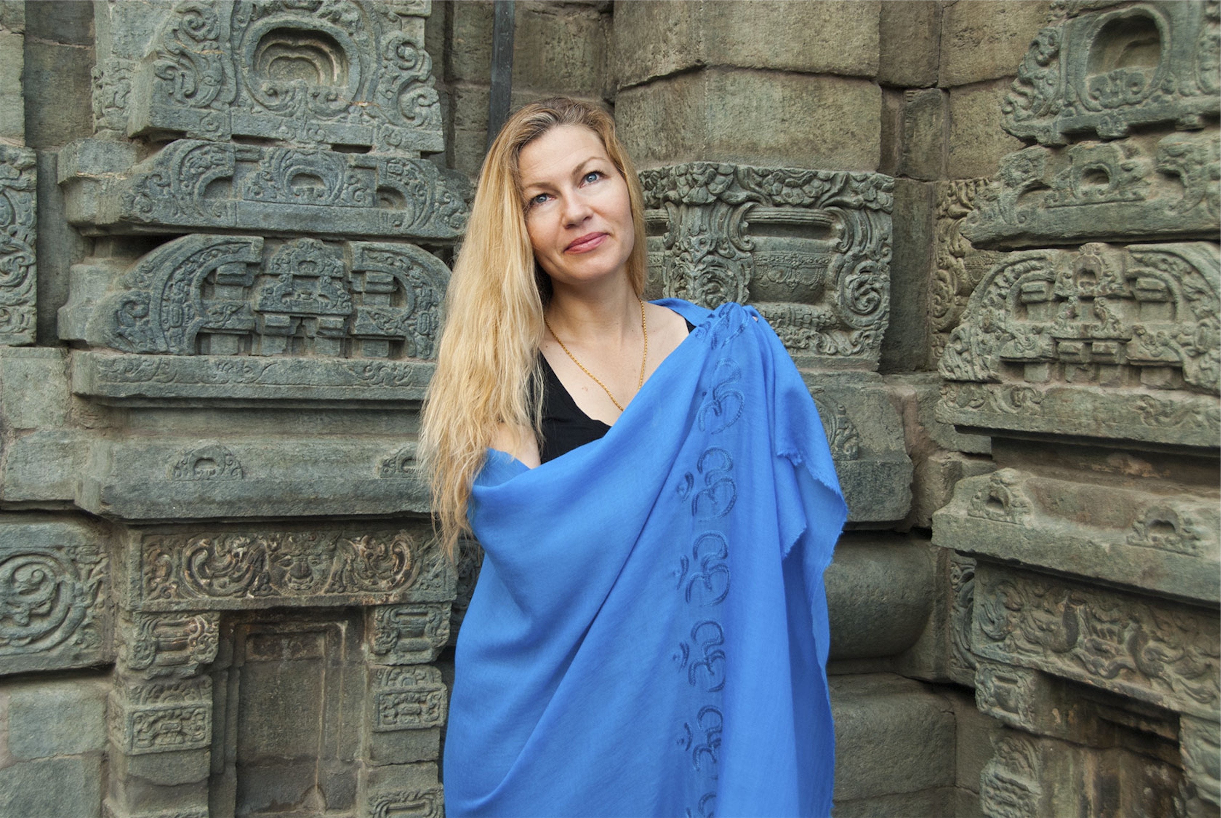 Blonde woman wearing a blue Shanti Om meditation shawl with Om symbols, standing against intricately carved stone walls of a Himalayan temple