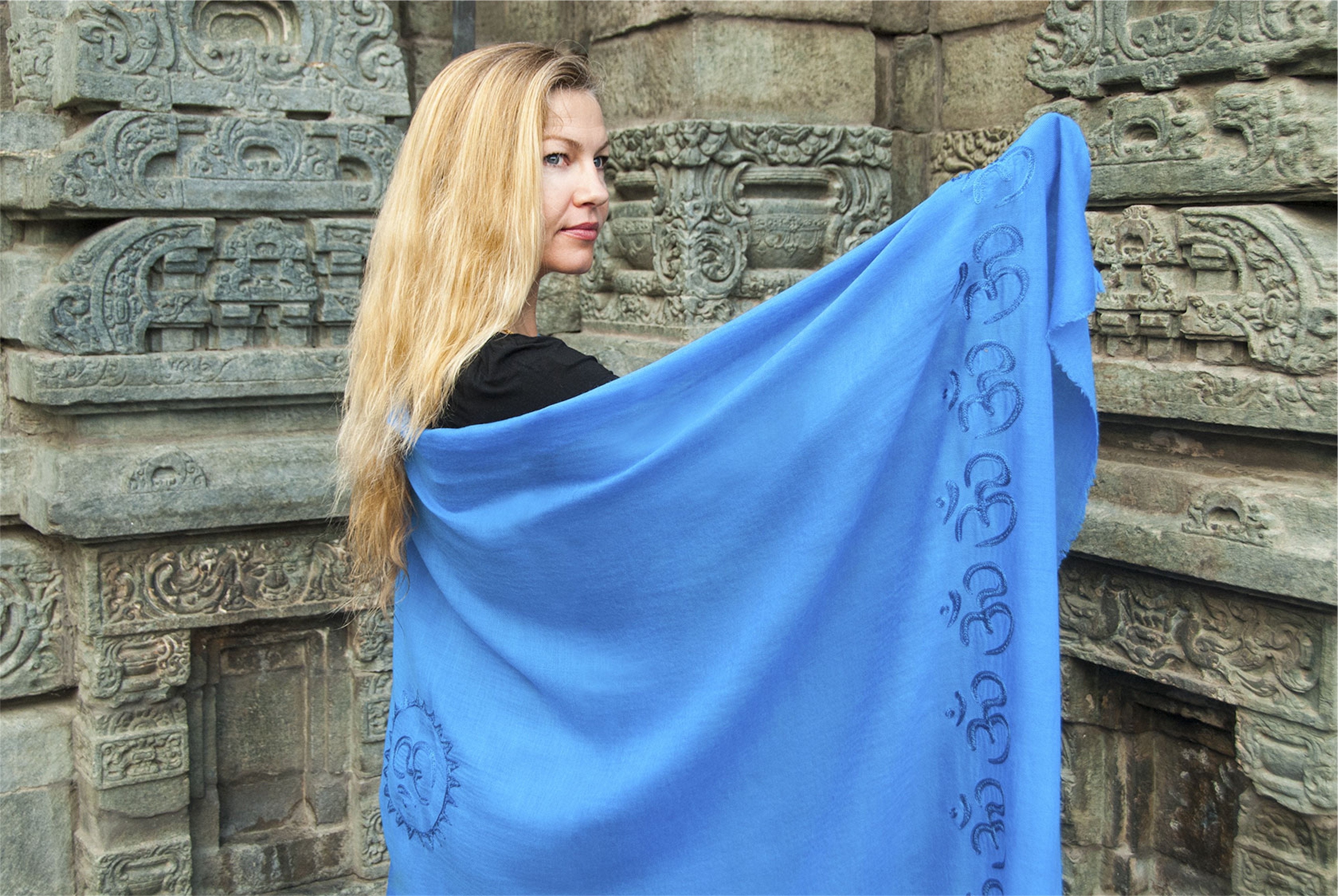 Blonde woman draped in a light blue Shanti Om meditation shawl with Om embroidery, standing near ancient Himalayan temple stone carvings