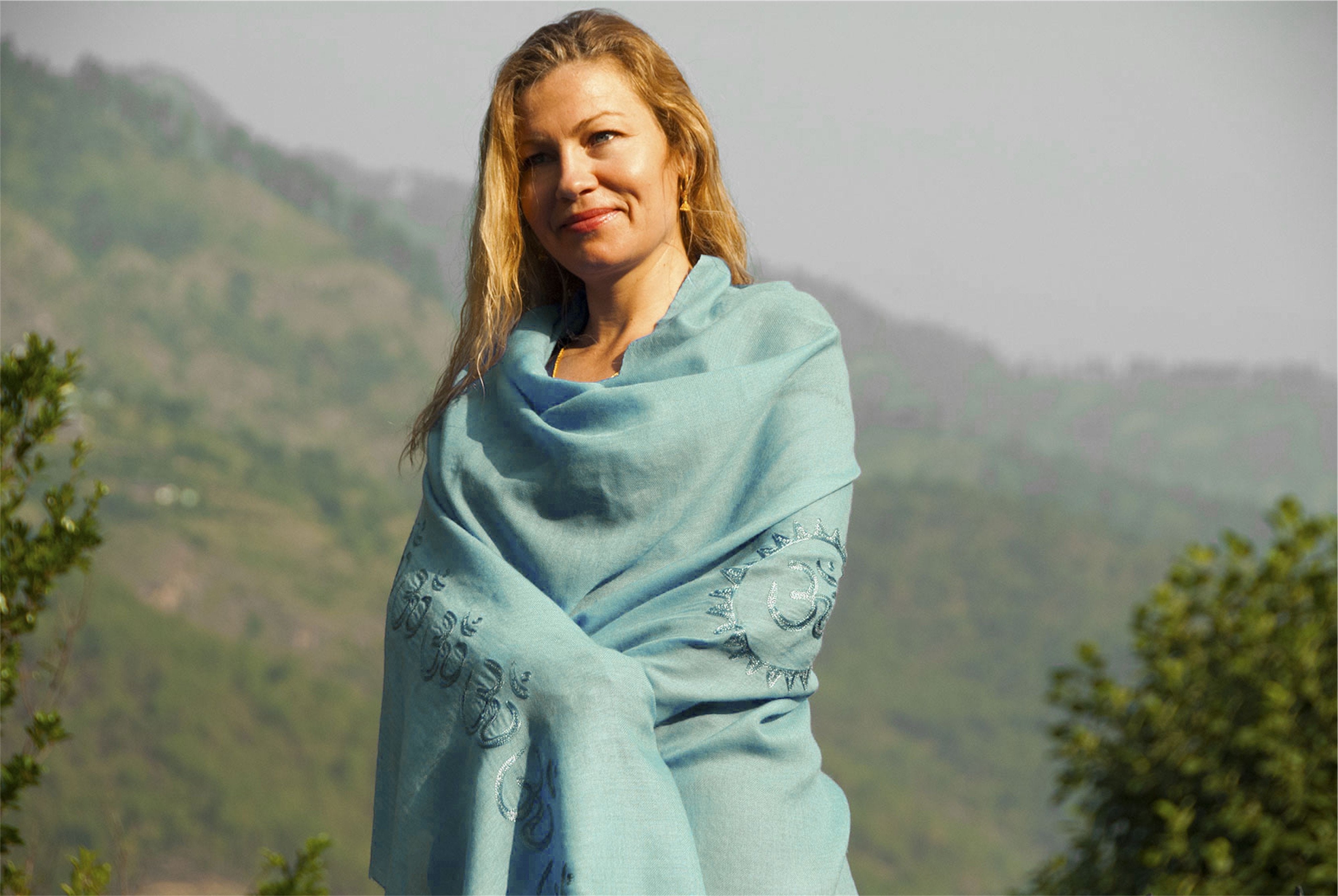 Woman wearing a light blue Shanti Om meditation shawl with intricate embroidery, standing against a backdrop of lush green mountains on a sunny day.