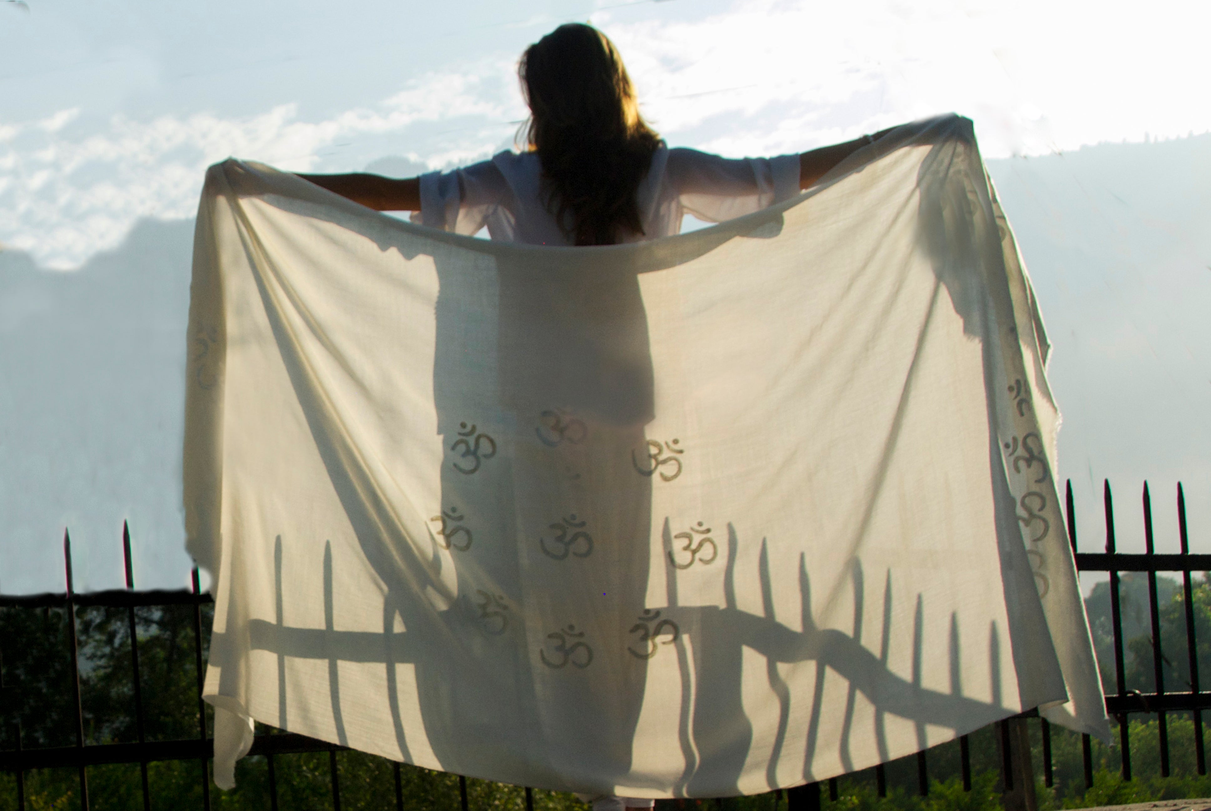 Backlit view of a person holding a cream Shiv Om Namah Shivaya meditation shawl with Om symbols, standing against a bright morning sky