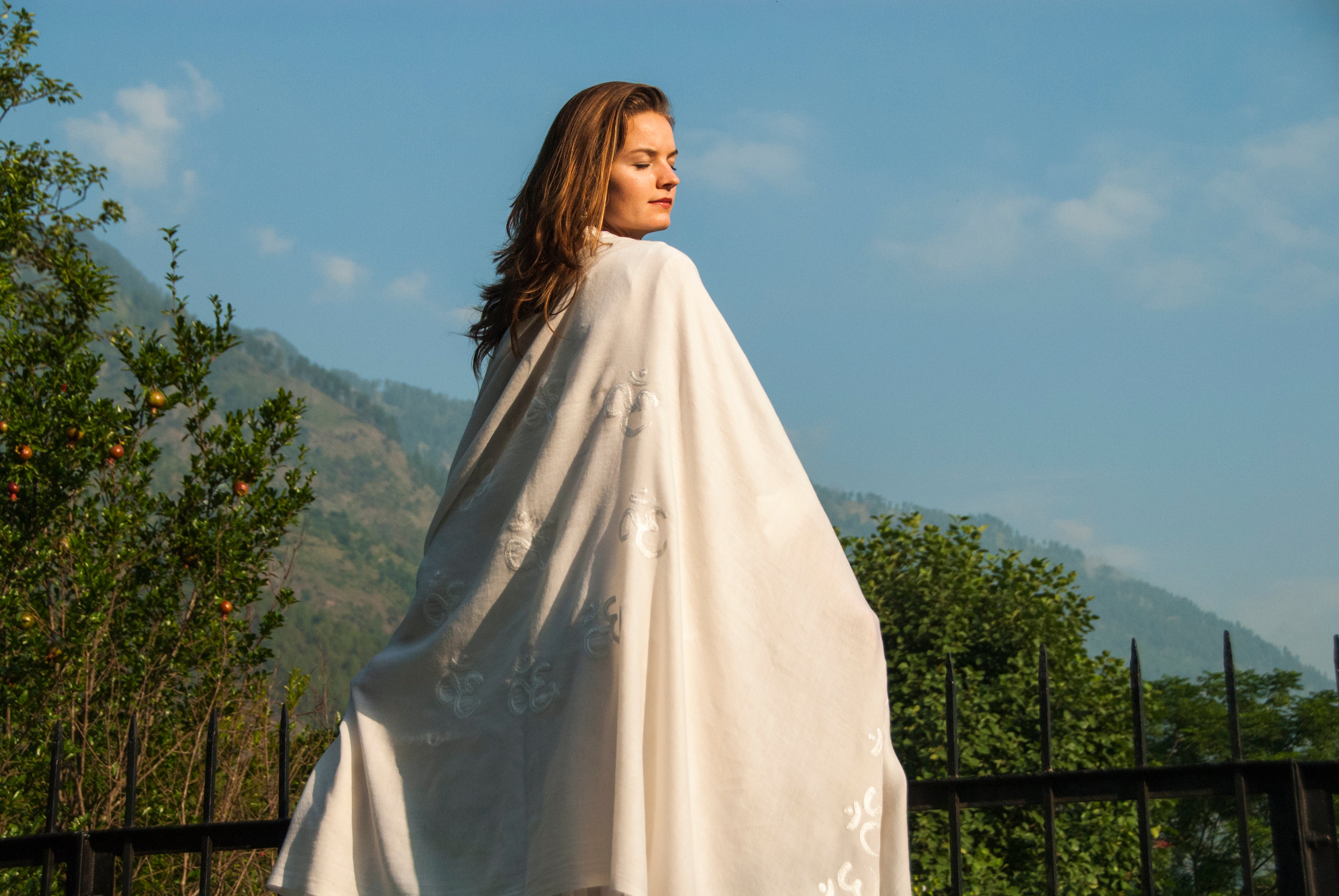 Portrait of a woman wearing a burgundy Shiv Om Namah Shivaya meditation shawl, seated in a vibrant garden with purple flowers in the background.