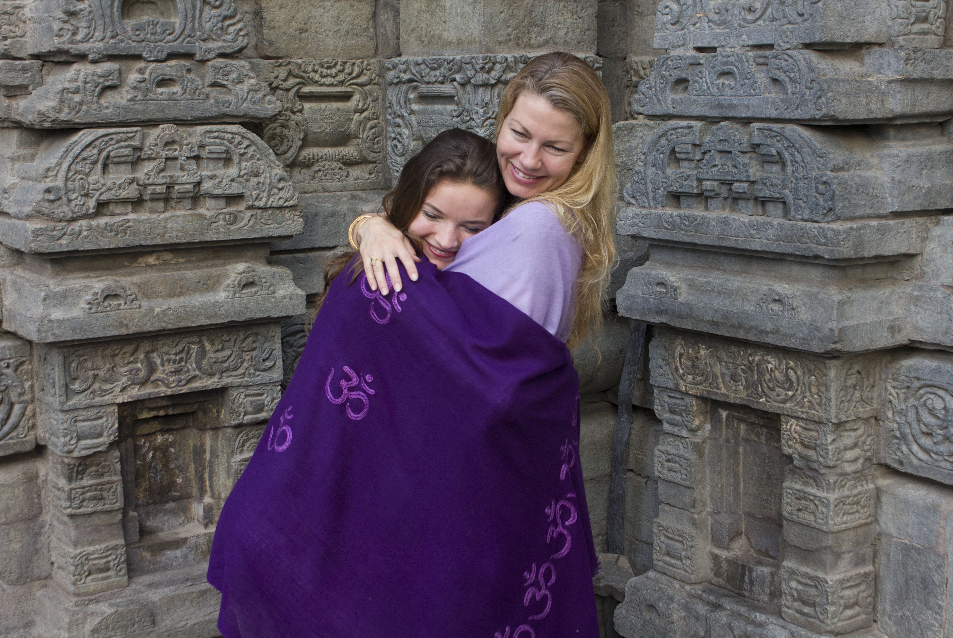 Two women wrapped in purple shawls embracing at a temple site.