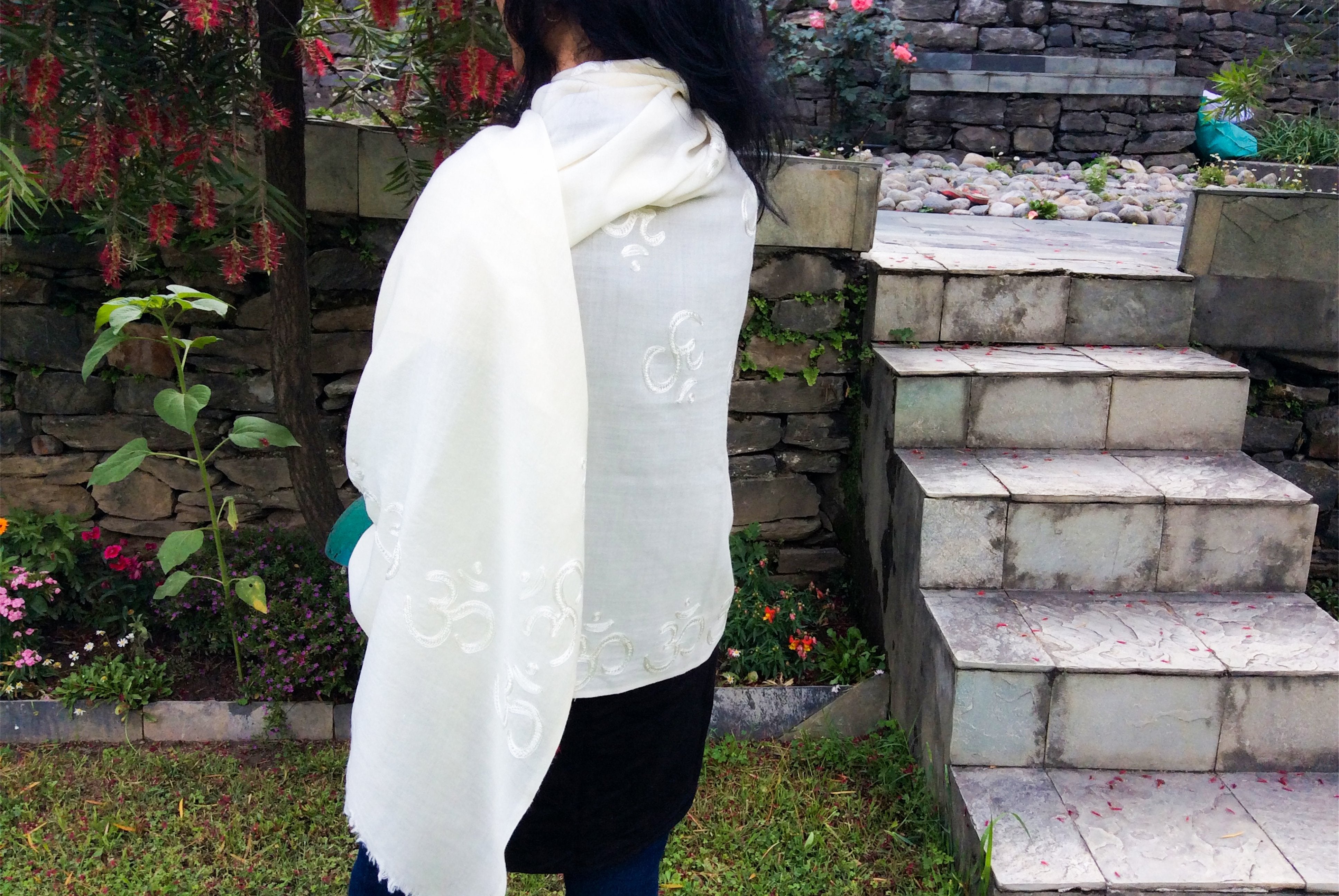 Woman wearing a white Om-embroidered yoga shawl in a peaceful garden setting, with stone steps and flowering plants in the background.