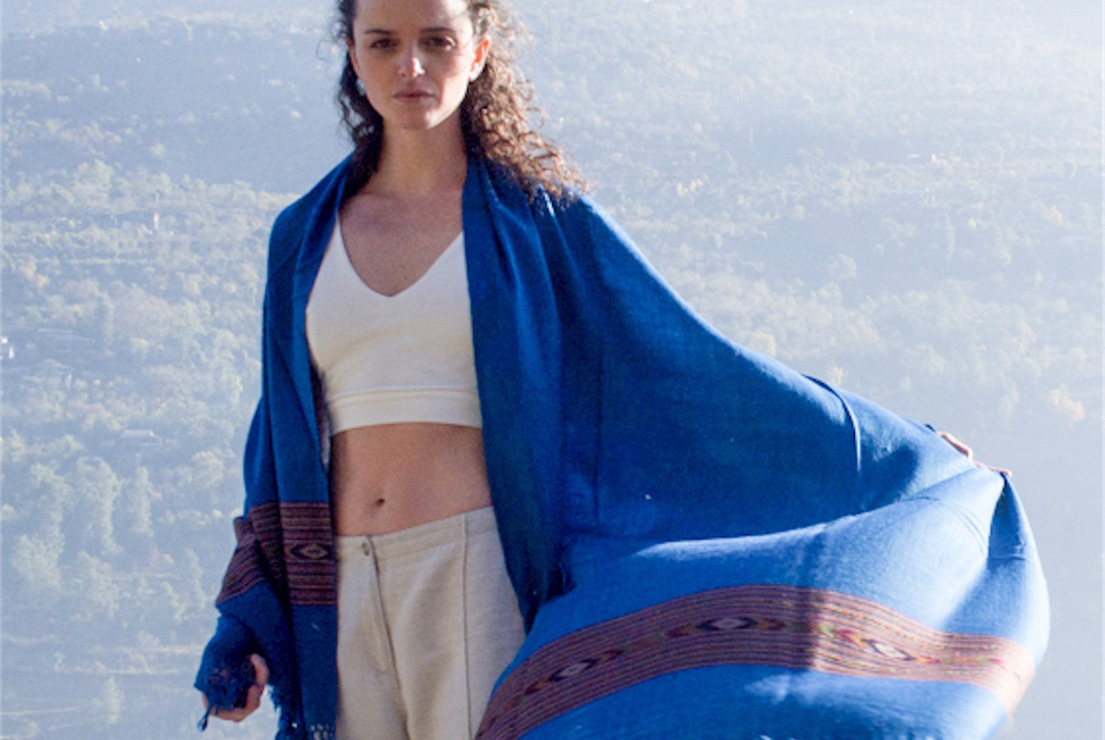 A woman wearing a blue shawl with an ethnic pattern with a scenic mountain landscape in the background.