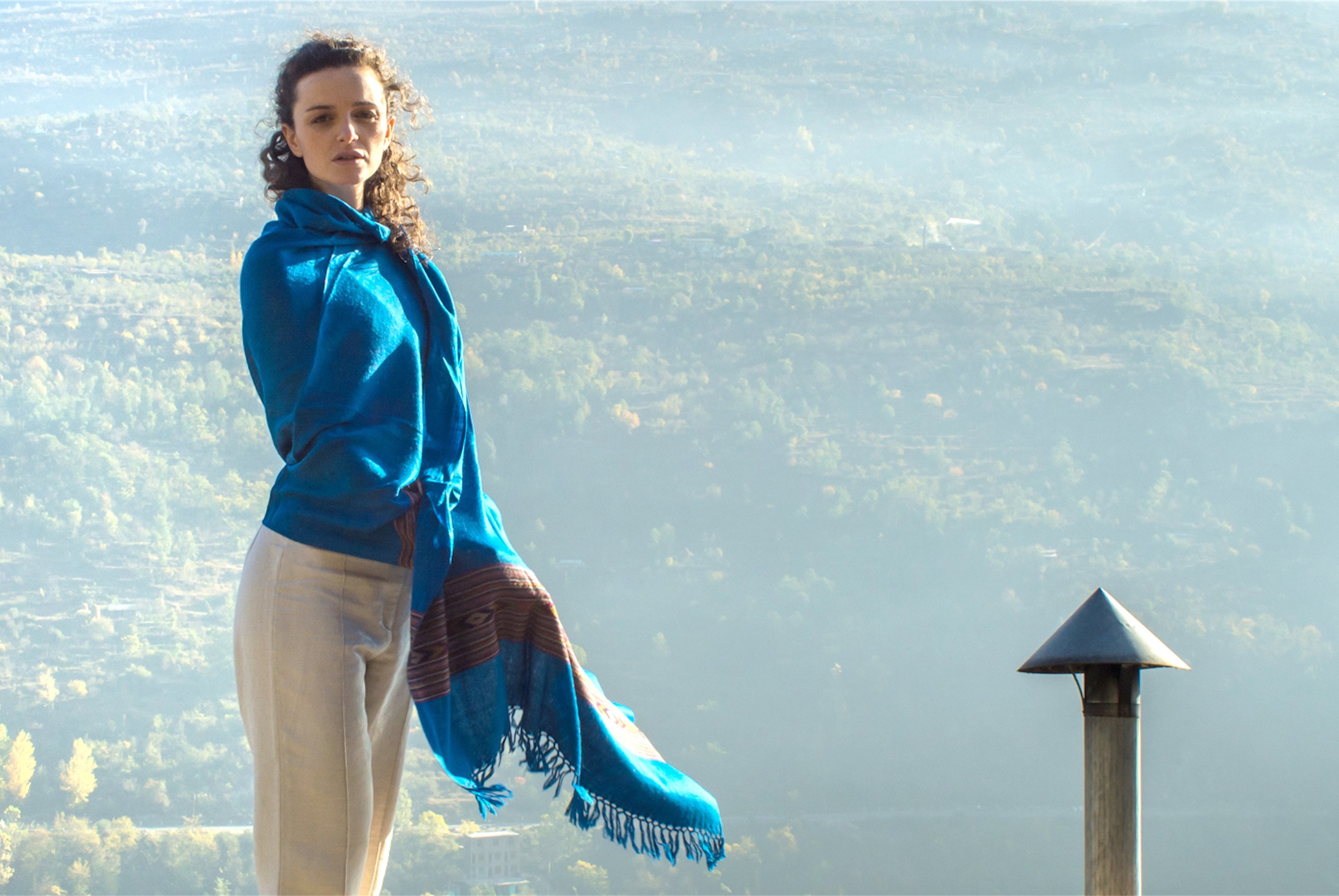 A woman wearing a blue shawl with an ethnic pattern, standing with a beautiful background of trees.