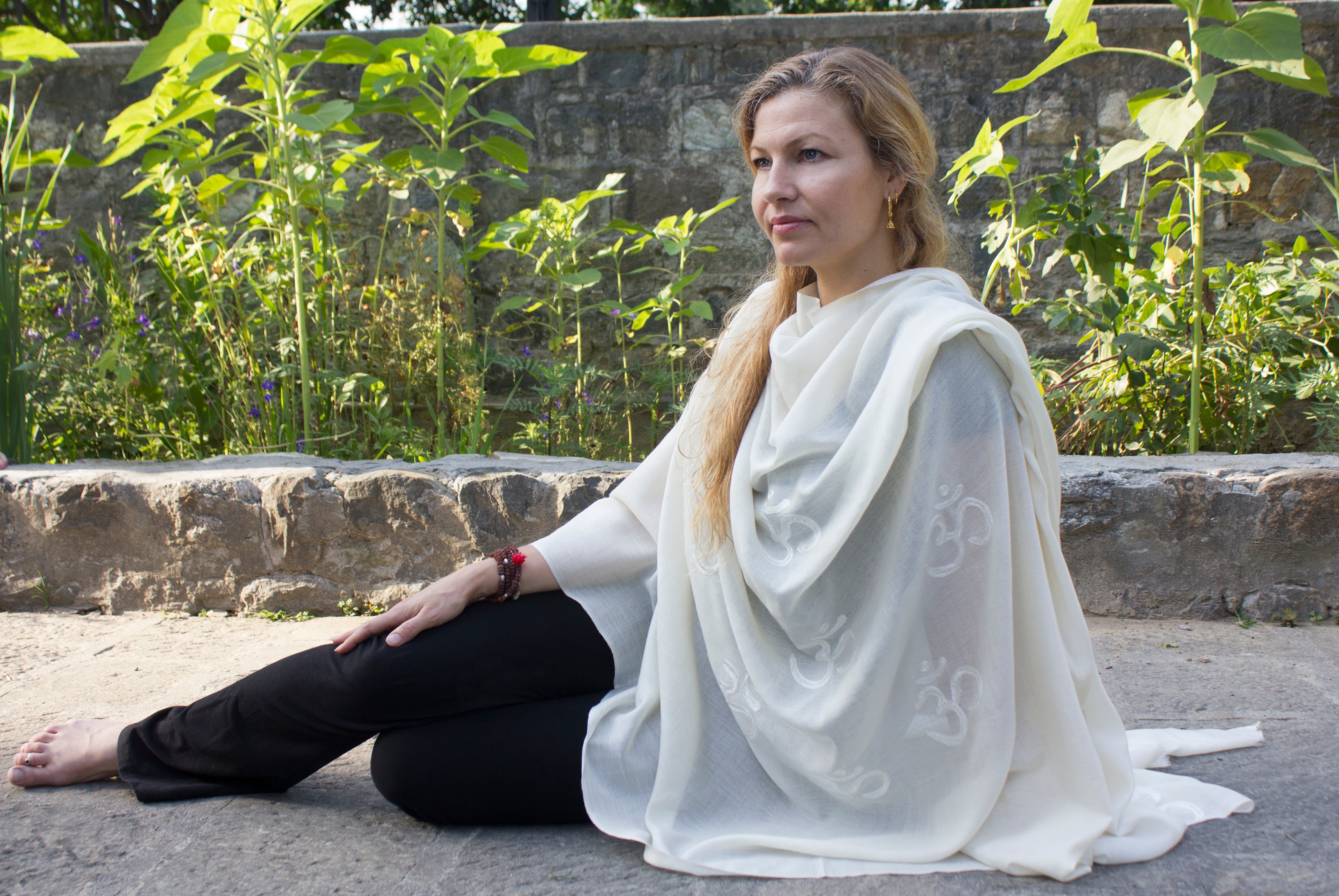 A woman seated on stone steps in a white shawl with Om symbols, surrounded by greenery and nature.