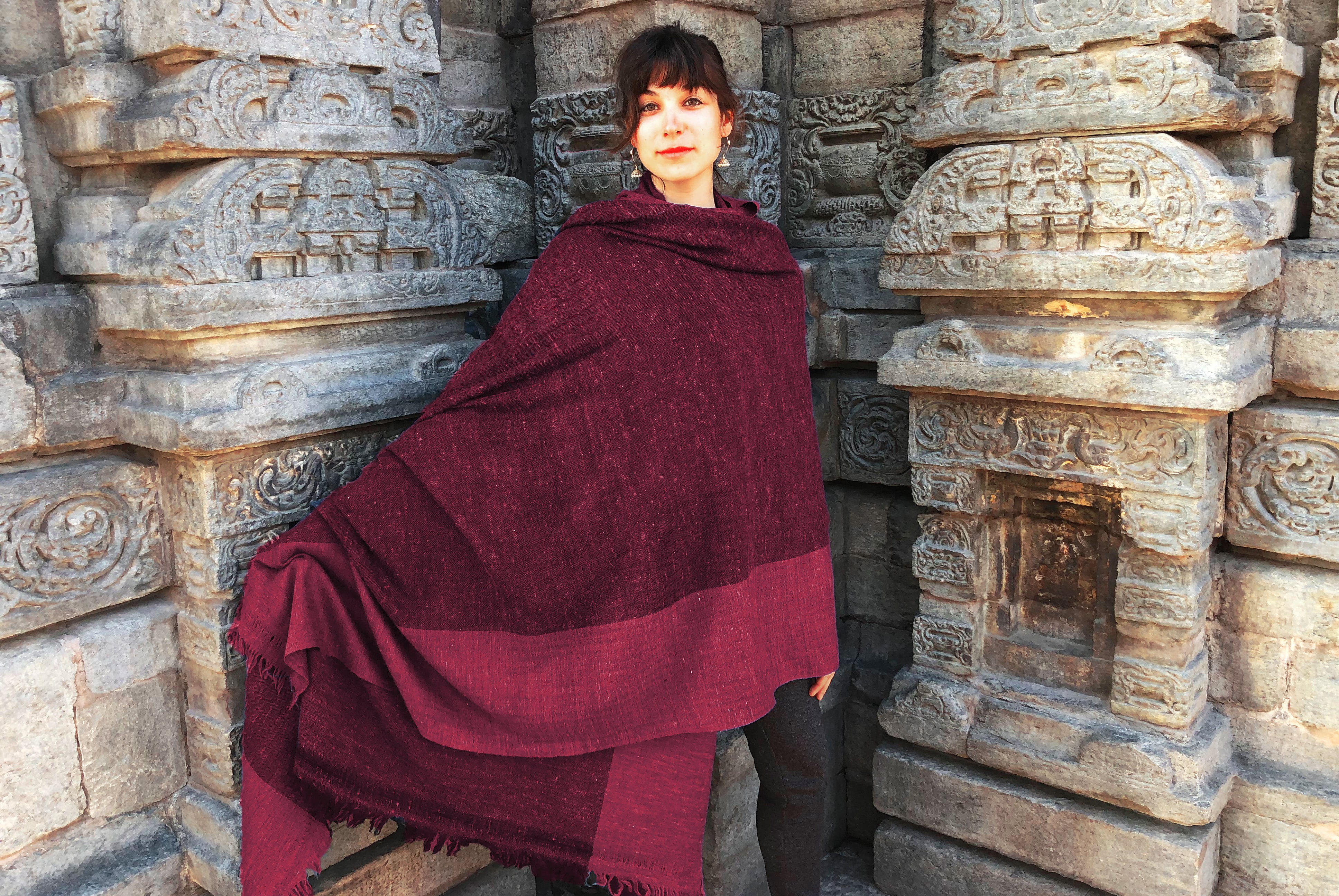 Woman wearing a burgundy shawl, standing by a stone-carved wall in a traditional setting.